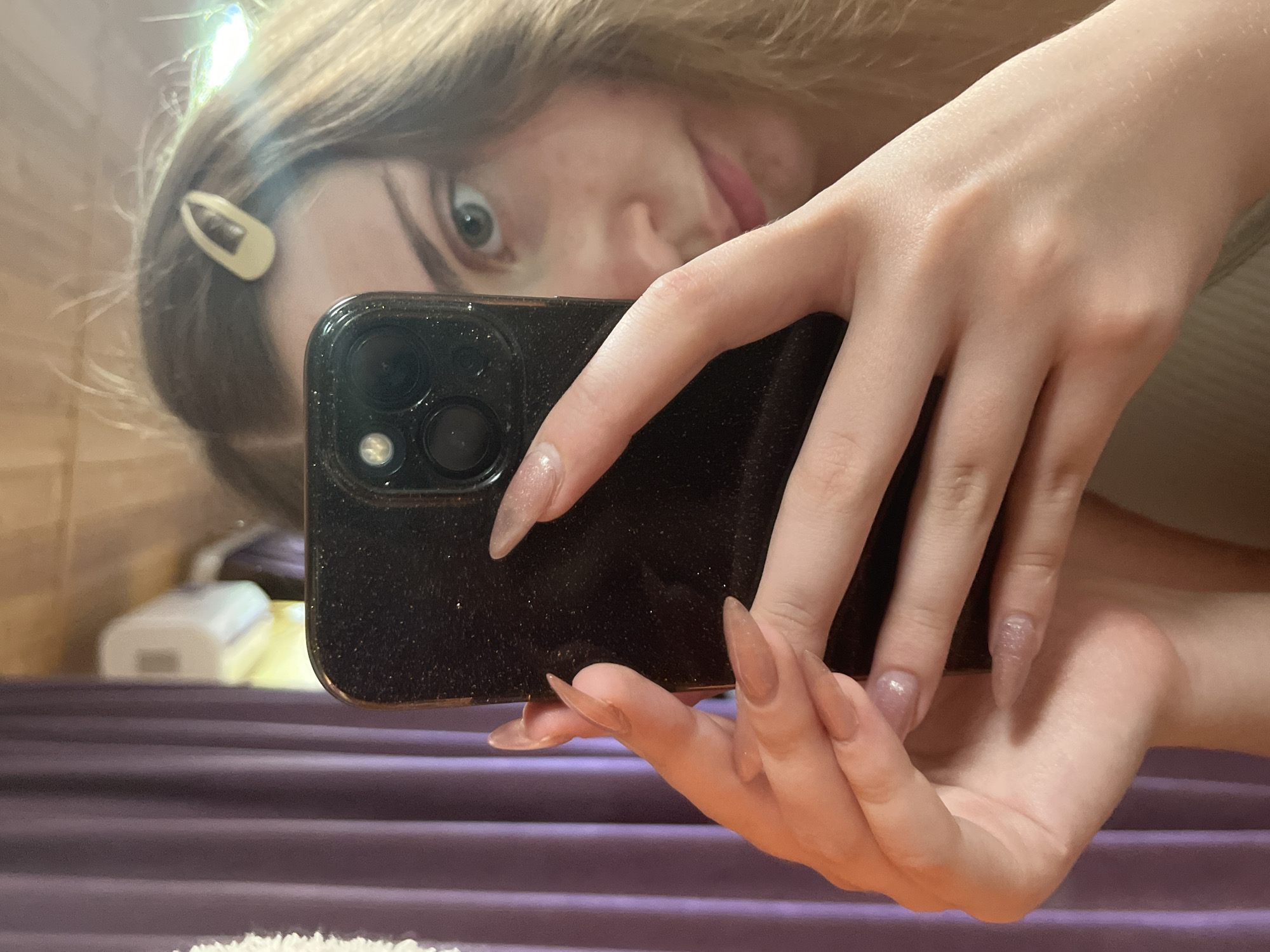 A young woman with light brown hair, held back by a beige hair clip, takes a mirror selfie. She is holding a dark-colored smartphone in her right hand, angled to capture her reflection. Her left hand is also visible, with long, light brown, pointed nails, resting on the phone. She has light-colored eyes and her face is partially obscured by the phone. The background appears to be an indoor setting, with a wooden wall and a purple curtain on the left. There is also a glimpse of a white object, possibly an air conditioner, in the background. The image does not provide any specific details that would allow for the identification of a location in Ashkadar, Russia.