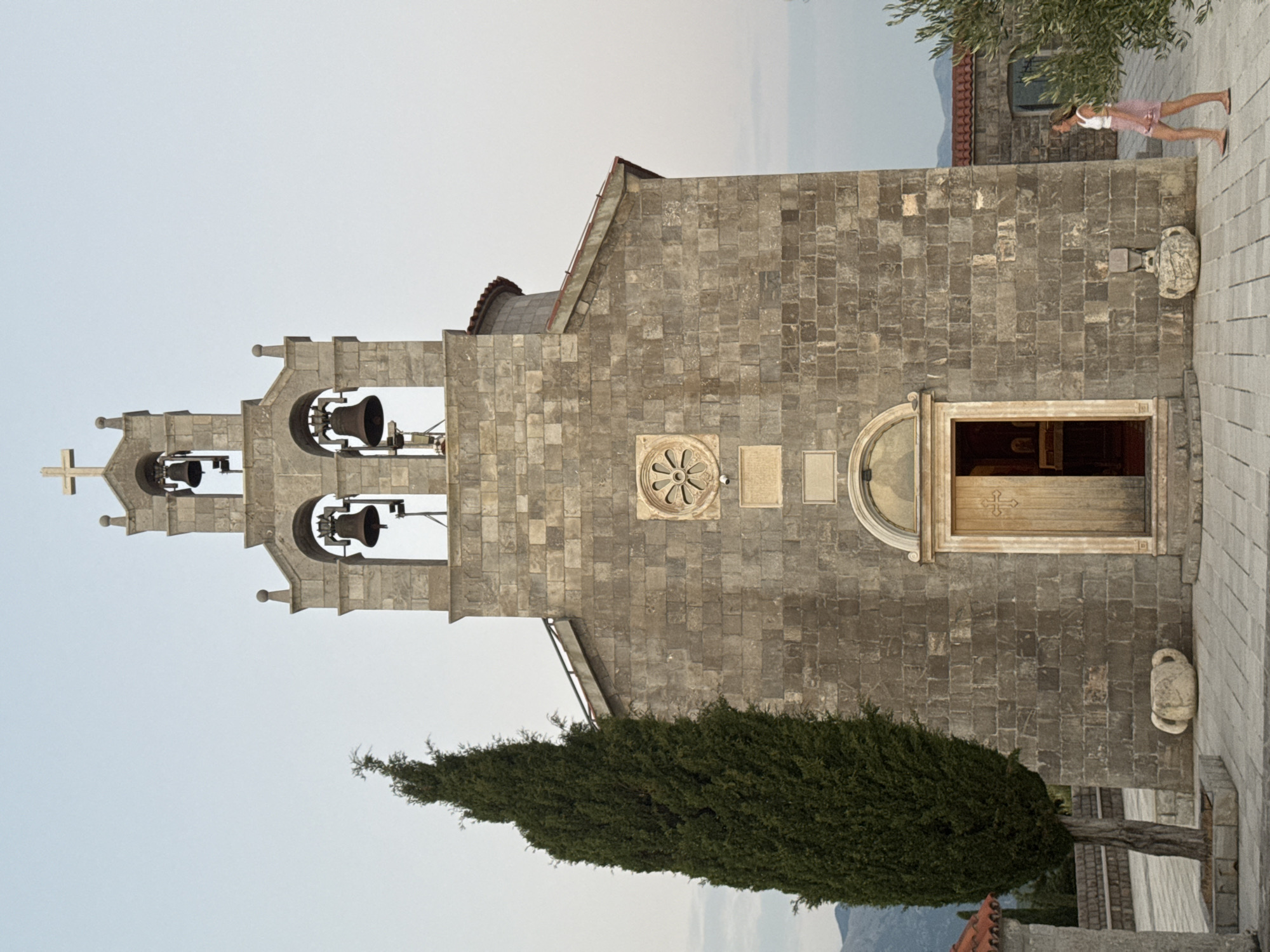 The image shows a stone church with a bell tower in Kovačko Polje, Montenegro. A lone woman in a white top and pink skirt walks on the paved area in front of the church. The church features a large wooden door with a cross design and a circular stone carving above it. The bell tower has two levels with arches, each housing a bell. A tall, slender evergreen tree stands to the left of the church. In the background, there are hills and other buildings, including one with a red tile roof. The sky is overcast and a pale blue.