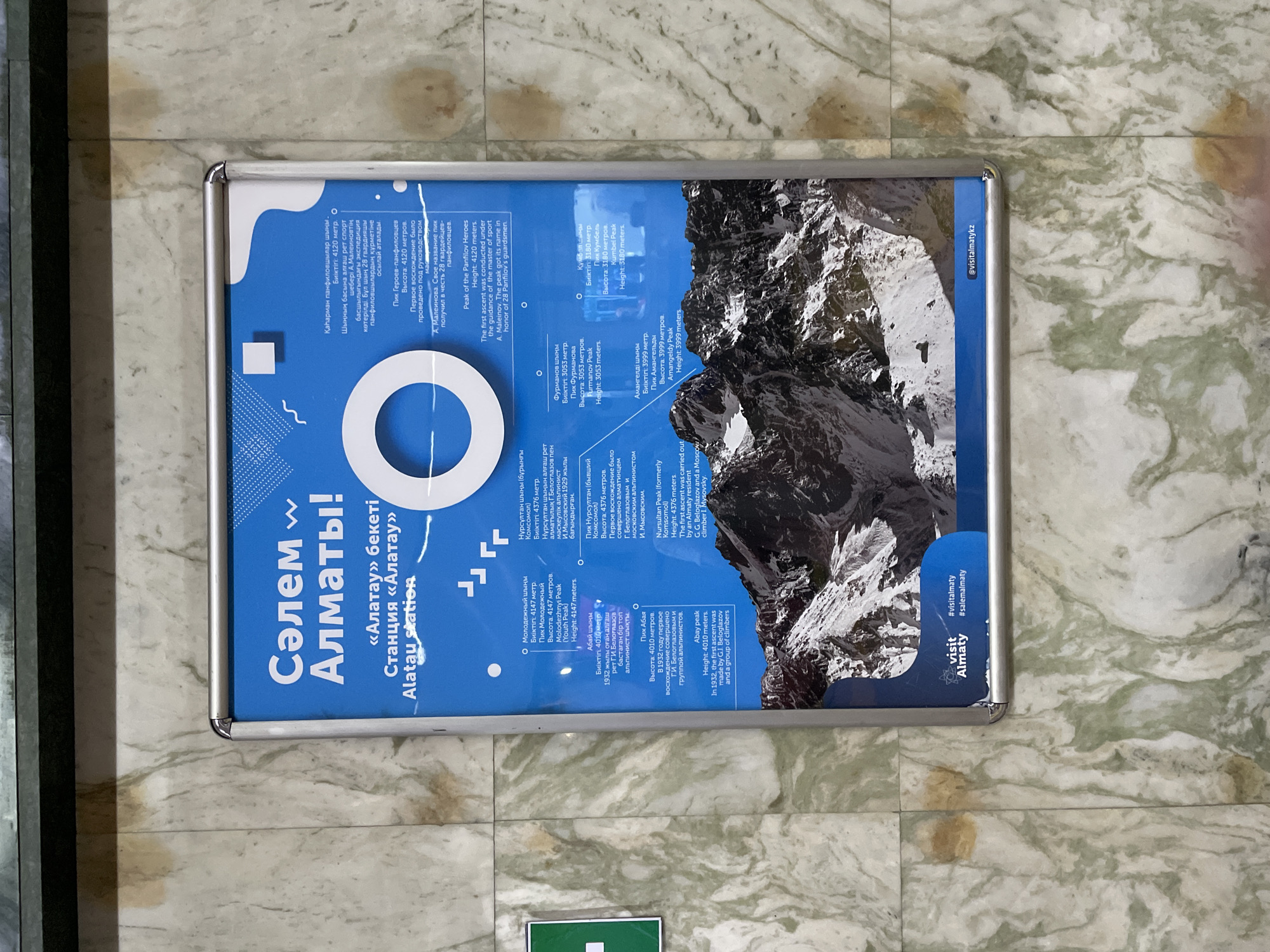 The image shows a large informational poster mounted on a marble-tiled wall. The poster is predominantly blue and features a mountain landscape at the bottom. The text on the poster is in Kazakh and Russian, with some English translation. The poster announces "Сәлем Алматы!" which translates to "Hello Almaty!" and specifically mentions "«Алатау» бекеті Станция «Алатау» Alatau station". It lists several mountain peaks, their heights, and some historical information, likely related to climbing expeditions. At the bottom of the poster are social media handles, #visitalmaty and #salemalmaly, and @visitalmatyky. The wall is a light-colored marble with green veining. The poster is framed in silver metal.