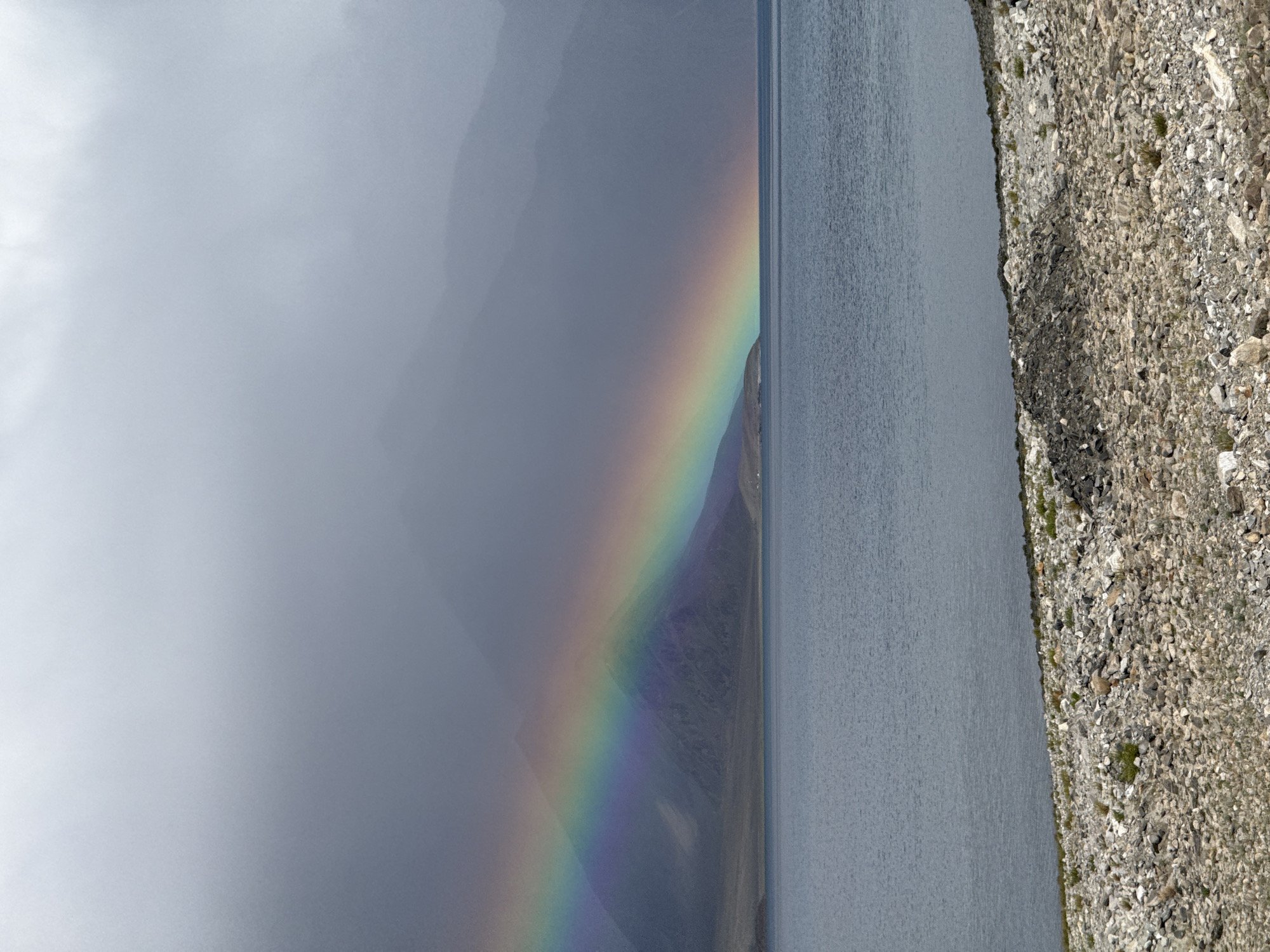 A rainbow arcs across a body of water with a mountainous landscape in the background under a cloudy sky. The foreground features a rocky shoreline. This is a scenic view of Pangong Tso lake in Man, India.