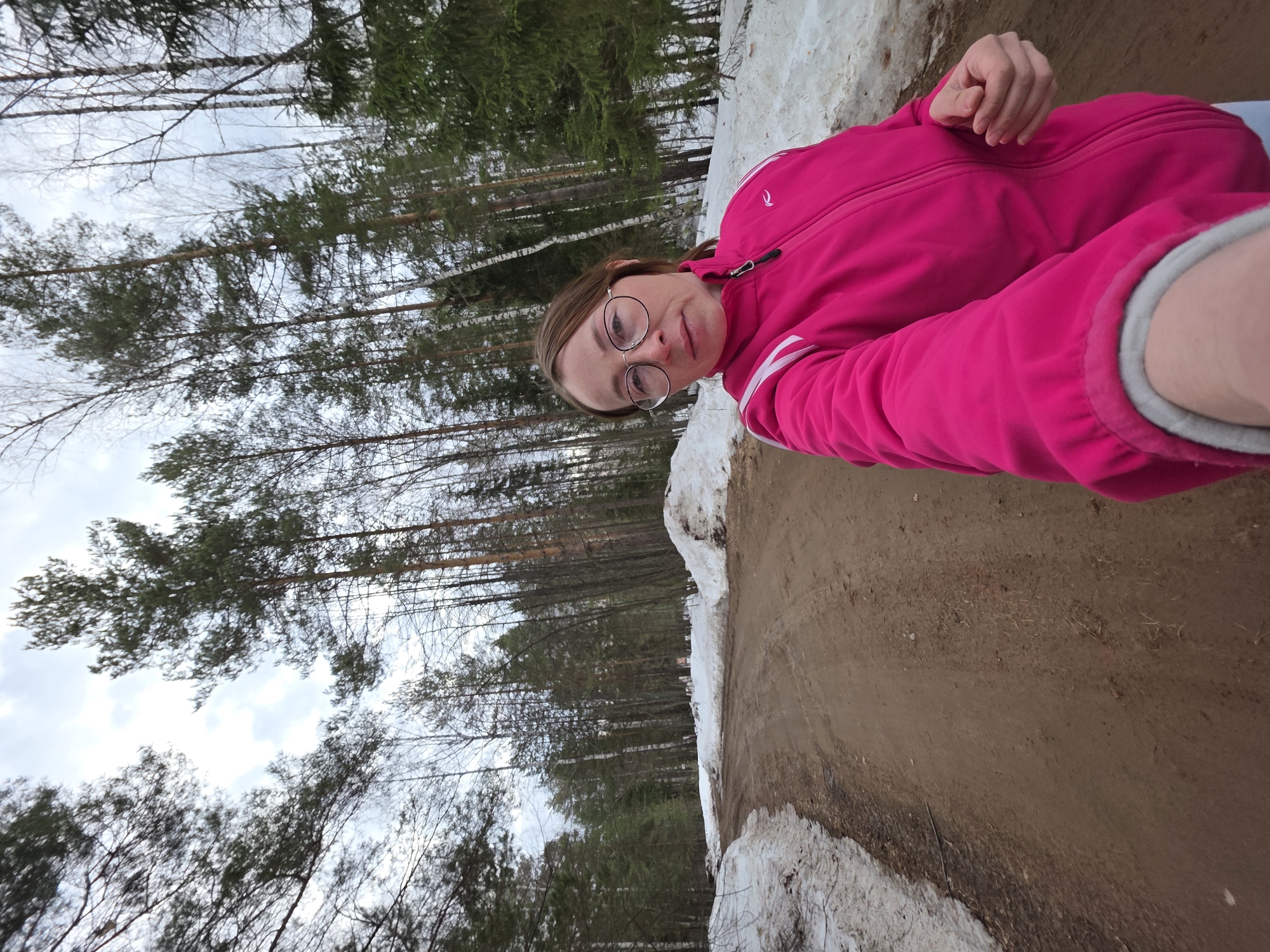 A person wearing glasses and a pink jacket is visible in the foreground. The person is positioned in front of a dirt path, which is bordered by snow and vegetation. In the background, there is a forest scene with trees of varying types under a partly cloudy sky. The location is in Novyi Urengoi, Russia.