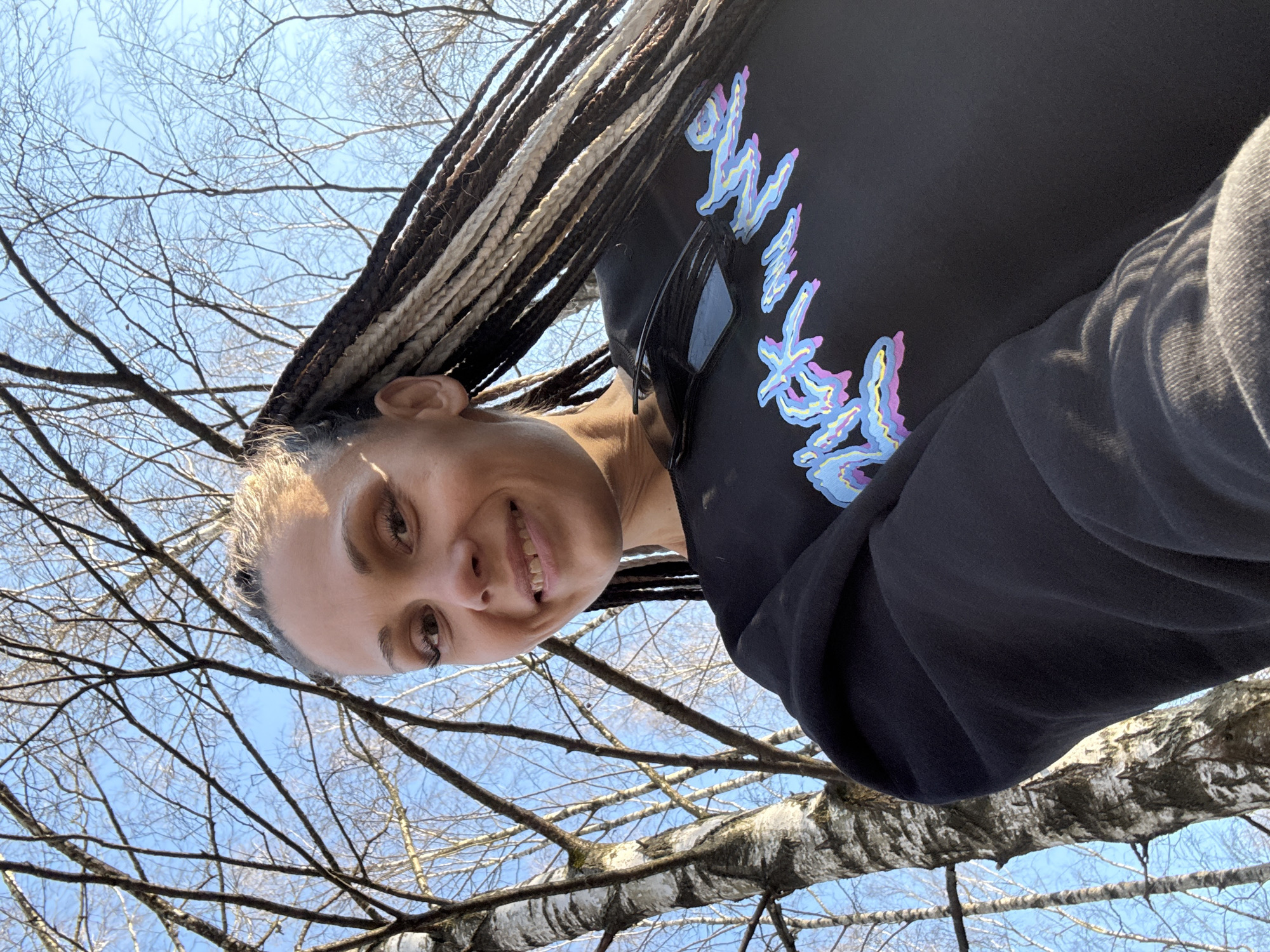 A low-angle shot captures a smiling individual with braided hair, partially light blonde, wearing a dark hoodie with a graphic design. The background features a leafless tree with white bark against a bright blue sky. The location is in a park, the city is Unknown, Unknown.