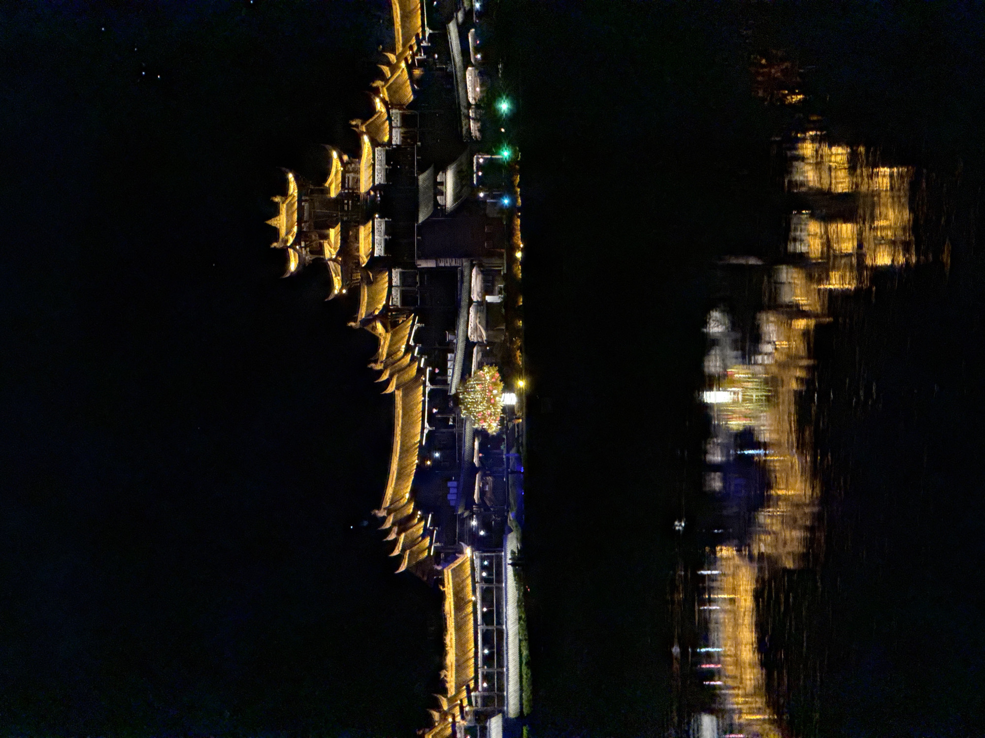 The image depicts a nighttime scene of a waterfront area in Zhuoshui, China. The photo is rotated 90 degrees clockwise.
A structure with traditional Chinese architectural elements is visible on the left side, illuminated with golden lights. There is a walkway and buildings with green and white lighting. The right side of the image shows a reflection of the waterfront area in the water. The water reflections appear blurred due to waves or movement. There are no people visible.