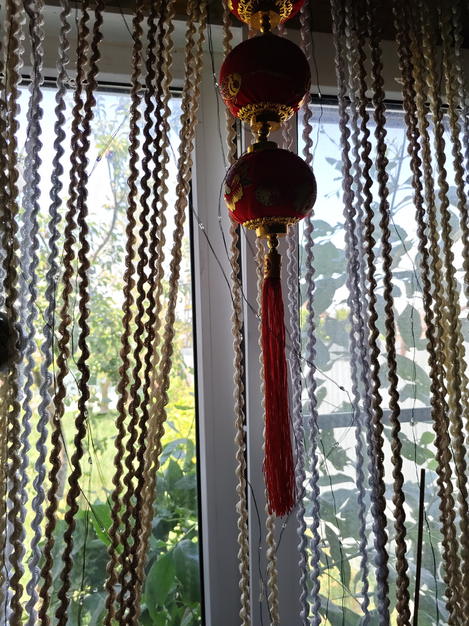 The image depicts a window with decorative curtains and two red Chinese lanterns hanging. The curtains are made of multiple strands of twisted material in various neutral colors, including beige, brown, and white, and appear to be hanging vertically. The Chinese lanterns are red with gold accents and a red tassel hanging from the bottom one. Through the window, greenery and foliage are visible, suggesting an outdoor environment. The scene appears to be indoors, with the window providing a view to the outside. No specific location within Rostov-on-Don, Russia is discernible from the image itself. There are no people or animals present. The overall lighting suggests daytime.