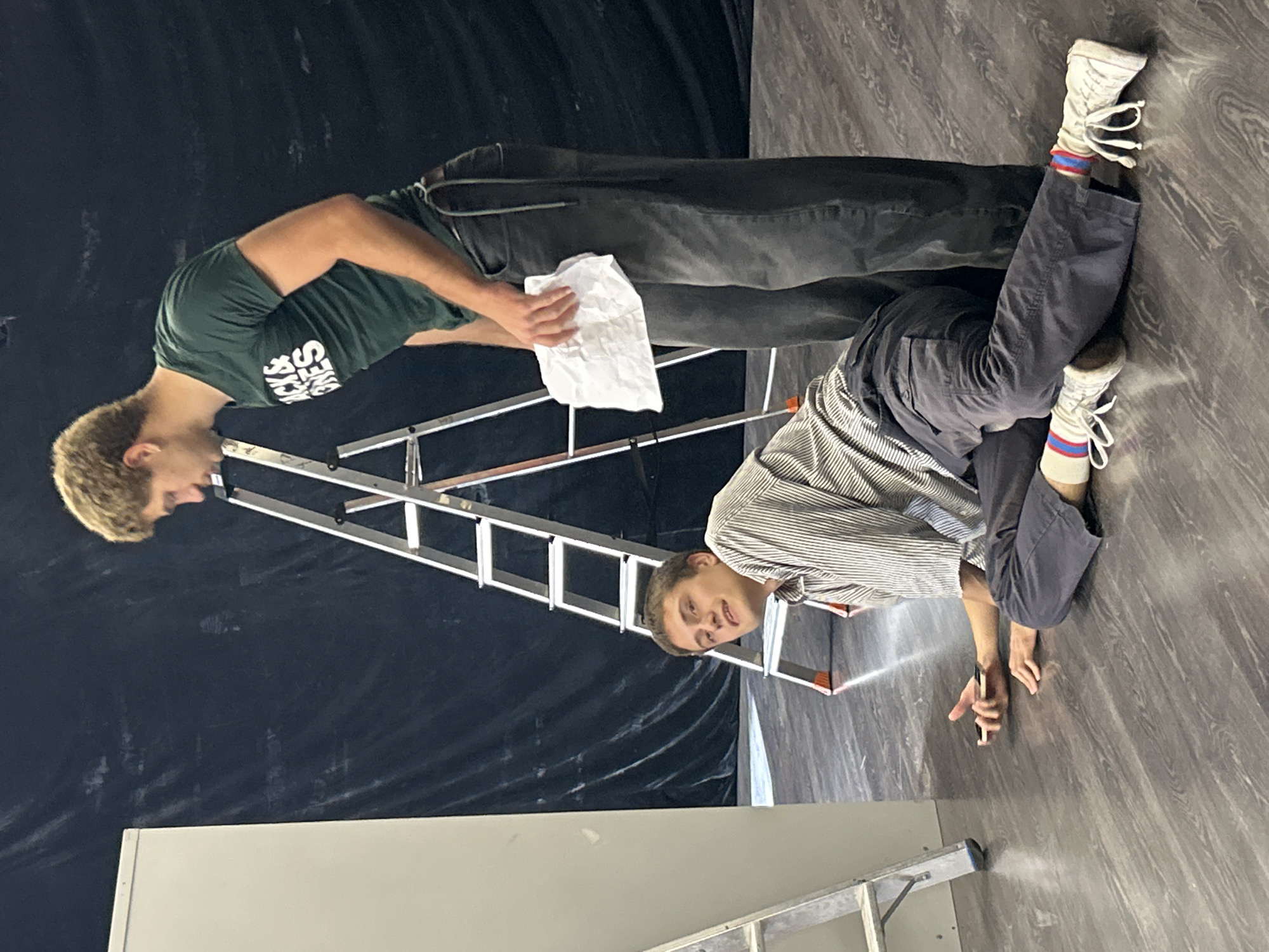 In an indoor environment with a dark, draped backdrop and a wooden floor, two individuals are present. A ladder is positioned centrally, with one person standing and the other lying on the floor.
The standing individual, a male with blond hair, wears a green t-shirt with "JACK & JONES" in white lettering and dark gray pants. He is holding a crumpled piece of white paper in his right hand and is looking downwards.
The individual on the floor is also male, with light brown hair, and is wearing a striped collared shirt, dark gray pants, and white high-top sneakers with red and blue striped socks. He is lying on his right side, looking towards the viewer, with his left arm extended and his hand holding a smartphone. His right arm is bent and resting on the floor. A second, partially visible ladder is to the left of the frame, and a tall, light-colored object, possibly a door or panel, is behind it. The lighting appears to be from overhead.