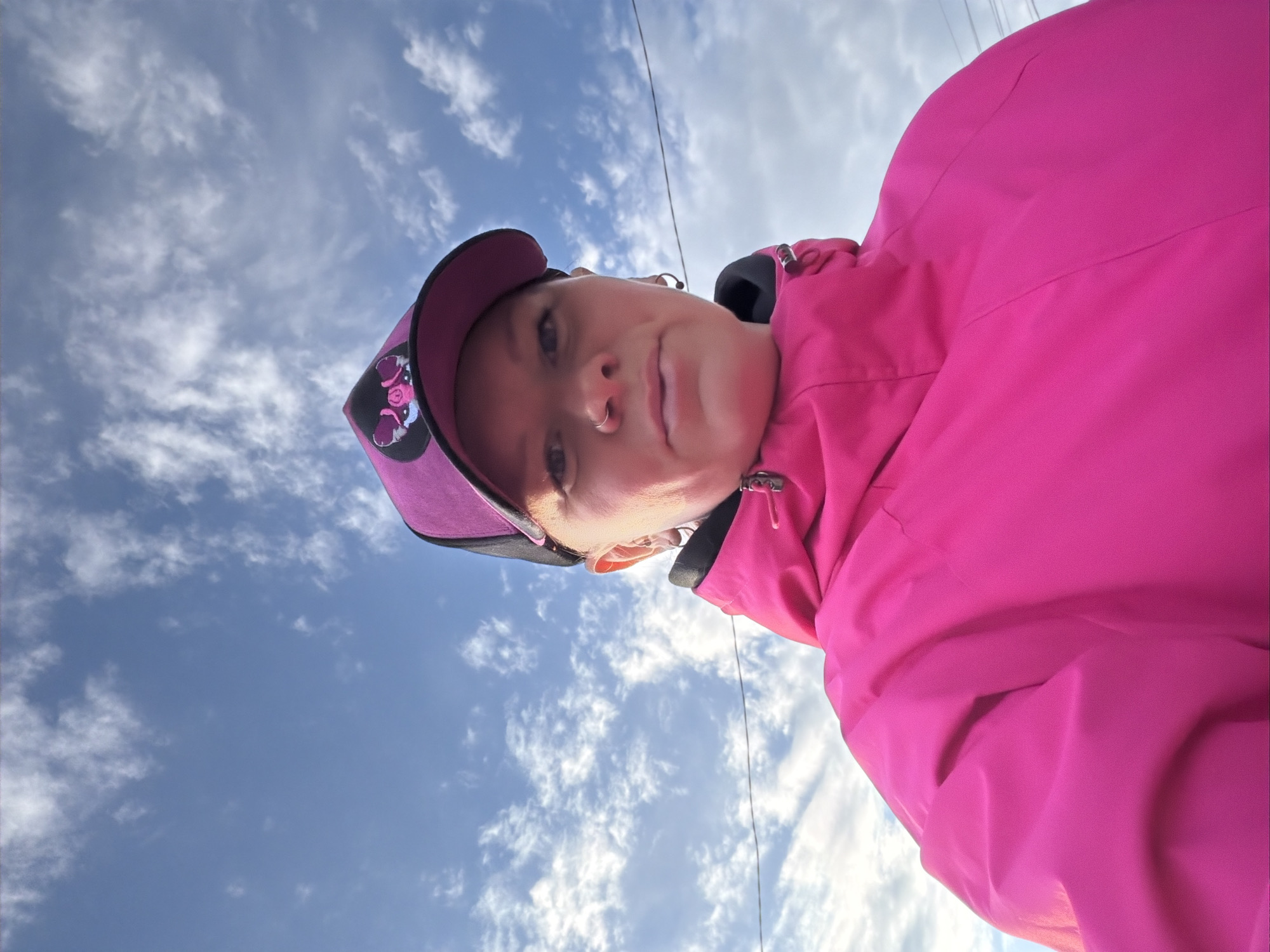 The image is a low-angle, rotated close-up of a person, likely a woman, taken against a bright blue sky with scattered white clouds. The subject is seen from the chest up, wearing a vibrant fuchsia-pink jacket and a purple baseball cap. The cap features a graphic design on the front, depicting what appear to be cartoon pigs or boars in pink and black. The person has a septum nose piercing and a neutral or slightly serious expression as they look directly into the camera. A thin, dark utility wire or cable is visible stretching across the sky in the background. The bright light suggests it is daytime with clear weather. The setting is an outdoor location in Chaikovsky, Russia. No visible text is clearly discernible.