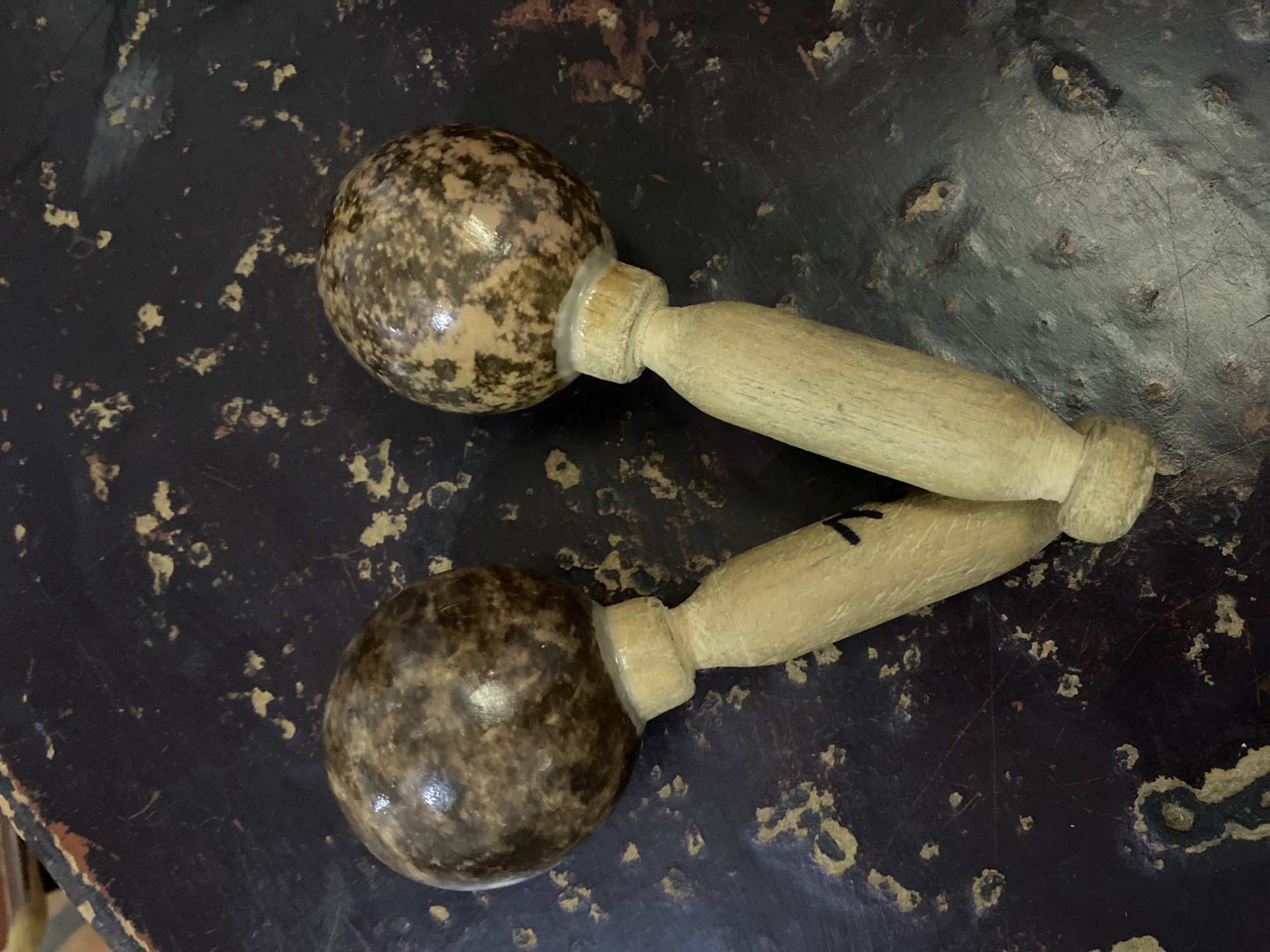 Two maracas are lying on a dark, textured surface. The maracas have spherical heads with a mottled brown and beige pattern, and wooden handles. The surface appears to be a table or countertop with worn paint and scuff marks. The lighting is overhead, creating some glare on the maracas' surfaces.
