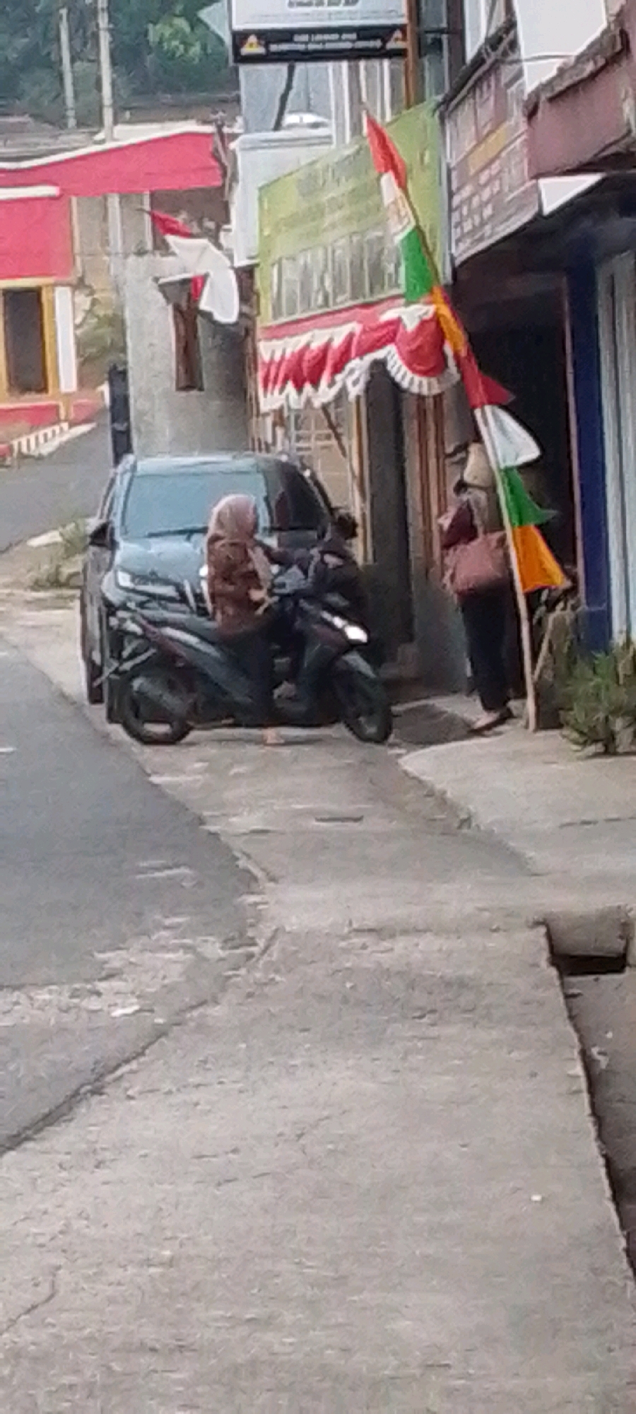 In Cilaku, Indonesia, a person wearing a hijab is seated on a dark-colored motorcycle, parked next to a dark-colored car. Another person stands to the right of the motorcycle and car, holding a bag. A flag, predominantly red and white with green and yellow accents, is planted nearby, extending upwards. The scene appears to be on a paved street with some damage and cracks, bordered by buildings. Red and white decorative bunting is visible above the doorway of one of the buildings. The overall impression is a street-level view of an urban setting.