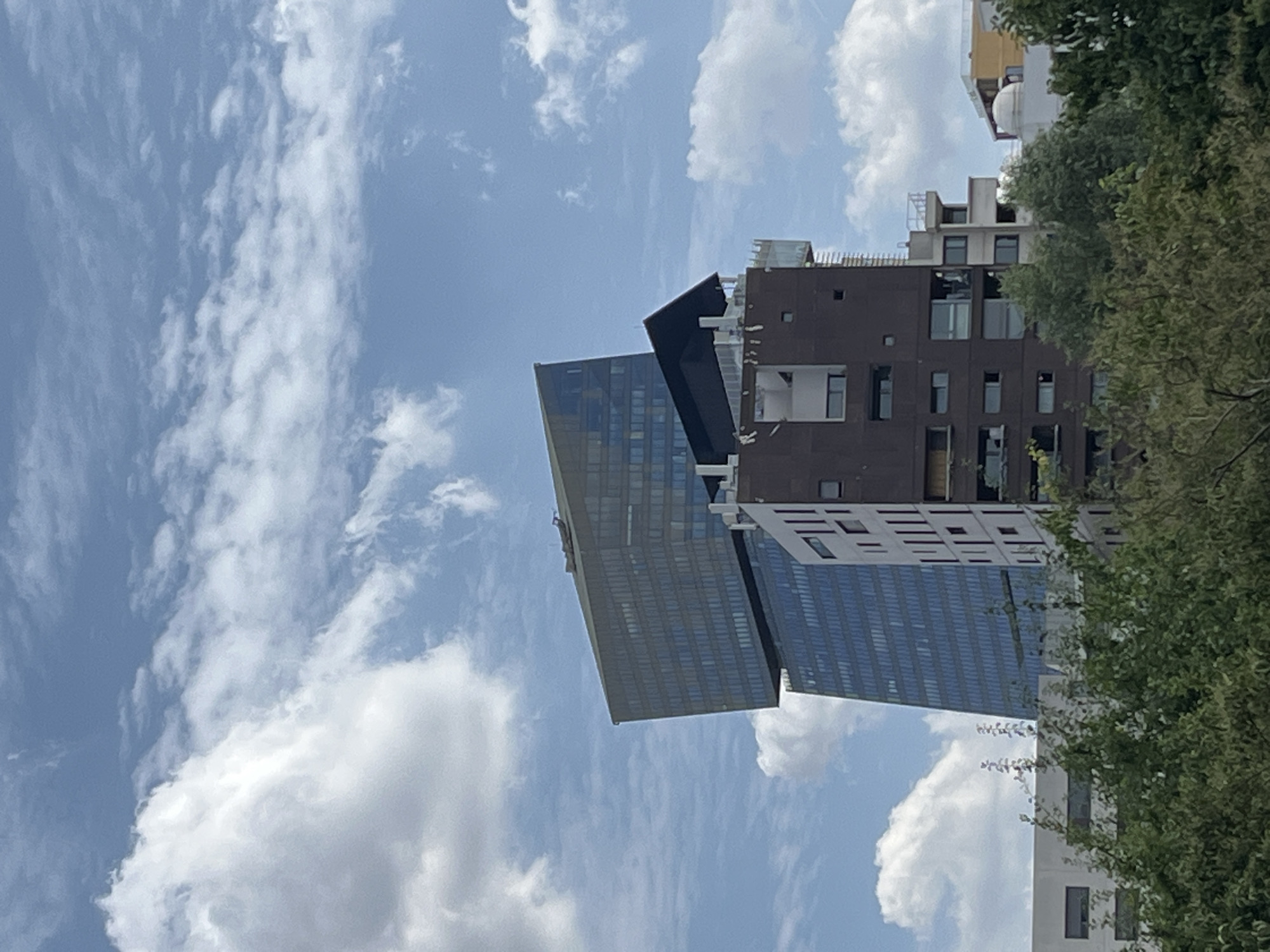 The image depicts a portion of modern architecture against a sky filled with white, puffy clouds. The primary subjects are two distinct buildings. One building is a sleek, glass-enclosed skyscraper with a distinctly angled, dark-colored section on its upper half. This glass skyscraper has a repeating pattern of windows or facade elements. Adjacent to it is another building with a darker, textured facade, featuring multiple windows, some appearing rectangular and others square. This building also has exposed structural elements and what appear to be balconies or outdoor spaces. Further in the background, glimpses of other structures are visible, including a building with a yellow facade. A dense green tree with visible branches and leaves partially obscures the right side of the frame, suggesting an outdoor setting. The sky is bright blue, indicating it is daytime. The clouds are scattered and appear illuminated by sunlight, suggesting fair weather. There are no visible people or discernable activity taking place. The overall impression is one of an urban or semi-urban environment with contemporary architectural design. The image is oriented vertically.