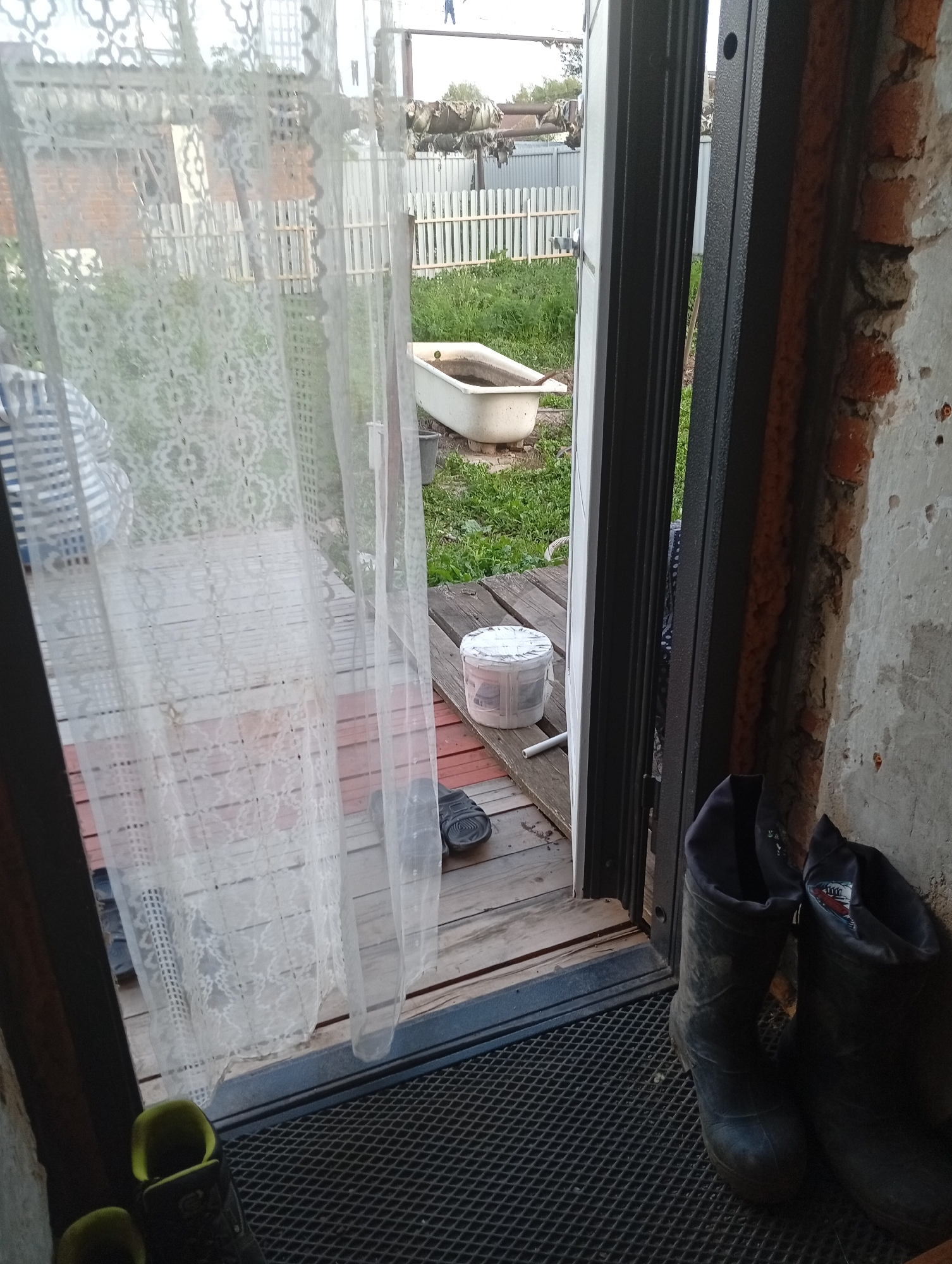 The image depicts an outdoor scene viewed from an entryway. Through a sheer white lace curtain, a bathtub is visible outdoors, placed on a wooden deck amidst overgrown grass. Next to the bathtub, a white bucket with a lid sits on the deck. A pair of black sandals are also present on the deck. The entryway itself has a grey metal door frame and a rough brick wall to the right. On the floor inside the entryway, a black rubber mat is laid out, with a pair of yellow and black work boots placed on it, and another pair of dark boots to the right. The background shows a white wooden fence and some structures. The overall impression is of a rustic, possibly rural, setting.