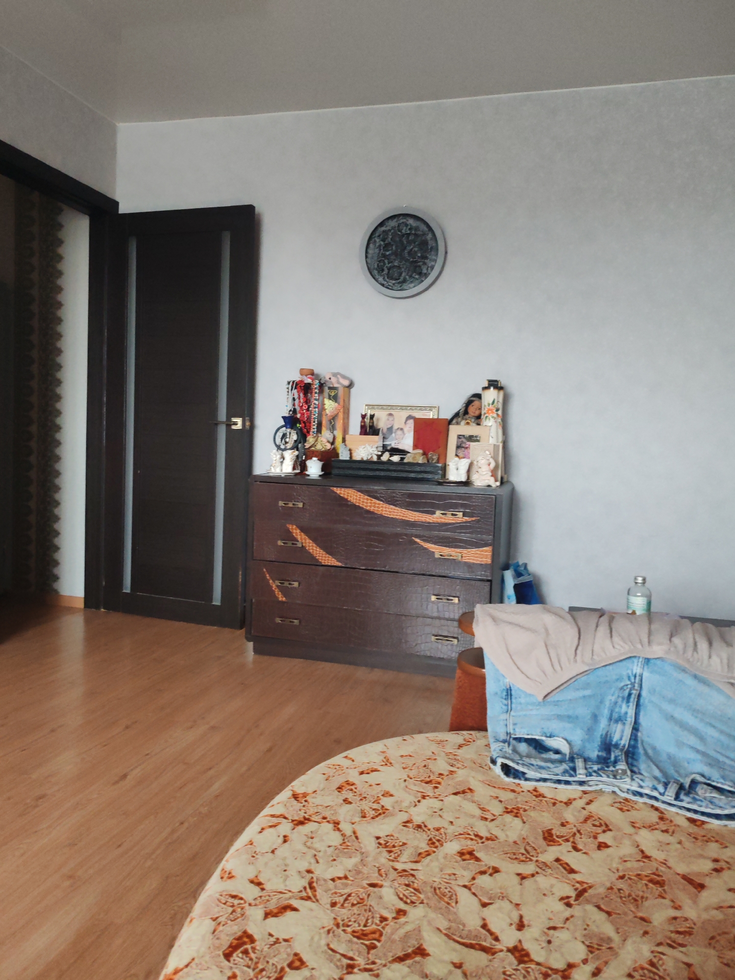 The image shows a room interior. A dark brown dresser stands against a light gray wall, on which hangs a round decorative object. Assorted items are placed on top of the dresser, including jewelry, figurines, and framed pictures. To the left of the dresser is a dark brown door with vertical light gray accents. In the foreground, a round table with a floral pattern is partially visible, with a tan shirt and a pair of blue jeans resting on it. The floor is made of light brown wood. There is no information about this being in Petropavlovsk-Kamchatsky, Russia, so it is unknown if that is the location. No people are visible.