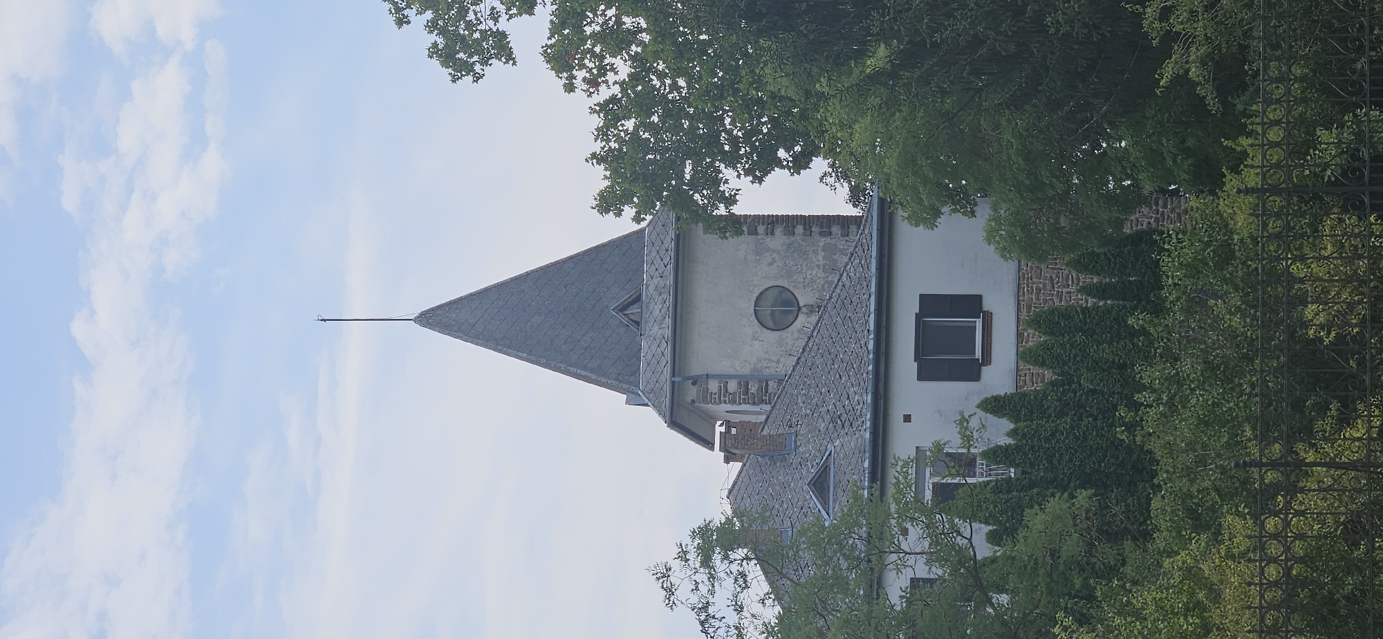 The image shows the upper portion of a building, likely a historic structure, with a prominent pointed tower and a steeply sloped roof covered in grey tiles. The tower is constructed with a mix of stone and grey plaster, featuring a small, circular window. A thin metal rod extends from the very apex of the tower. Below the tower, the building has white walls and a similarly tiled roof. A darker-colored louvered window is visible on the main body of the building. The structure is partially obscured by dense green foliage from various trees and bushes. A black, ornate metal fence runs along the bottom of the frame, separating the foreground from the building and vegetation. The sky above is light blue with scattered white clouds. This structure is the historical building of the former Roman Catholic parish church in Hévíz, Hungary.