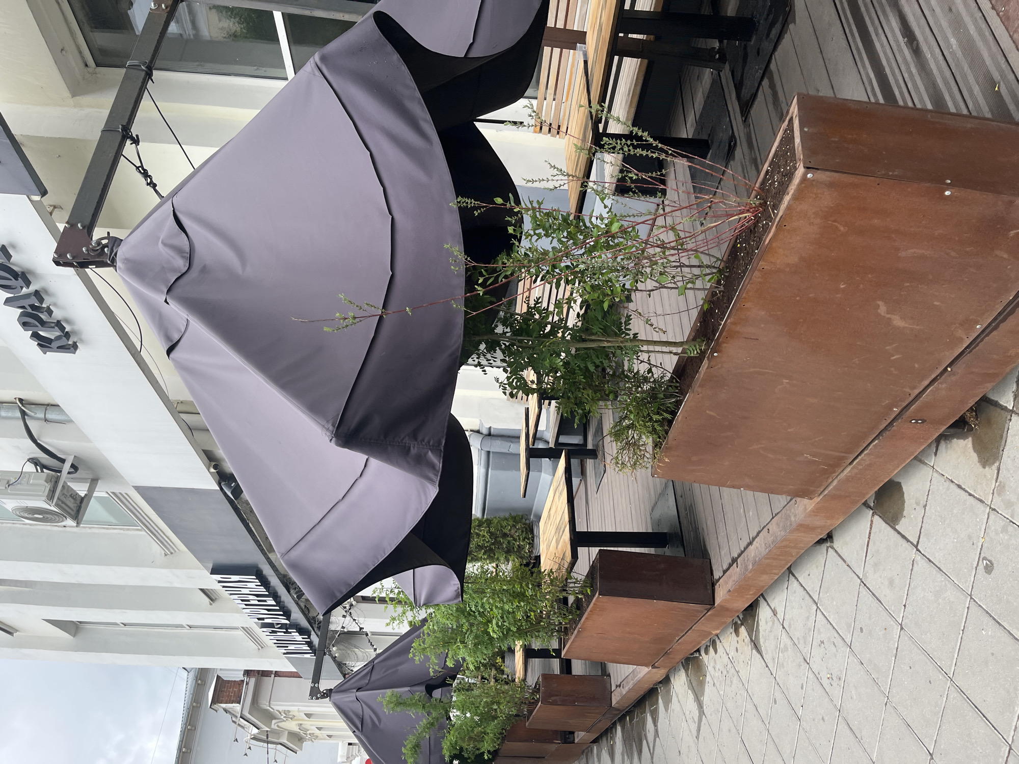 The image shows an outdoor seating area of a restaurant or cafe in an unknown city. The seating consists of wooden tables and benches, protected from the elements by large grey umbrellas. The area is decorated with long, rectangular metal planters filled with greenery. The floor is tiled and appears wet, possibly from recent rain. The restaurant's signage is visible, but the name is not fully legible. The building facade is white with dark trim. Overall, the scene suggests a typical urban outdoor dining space.