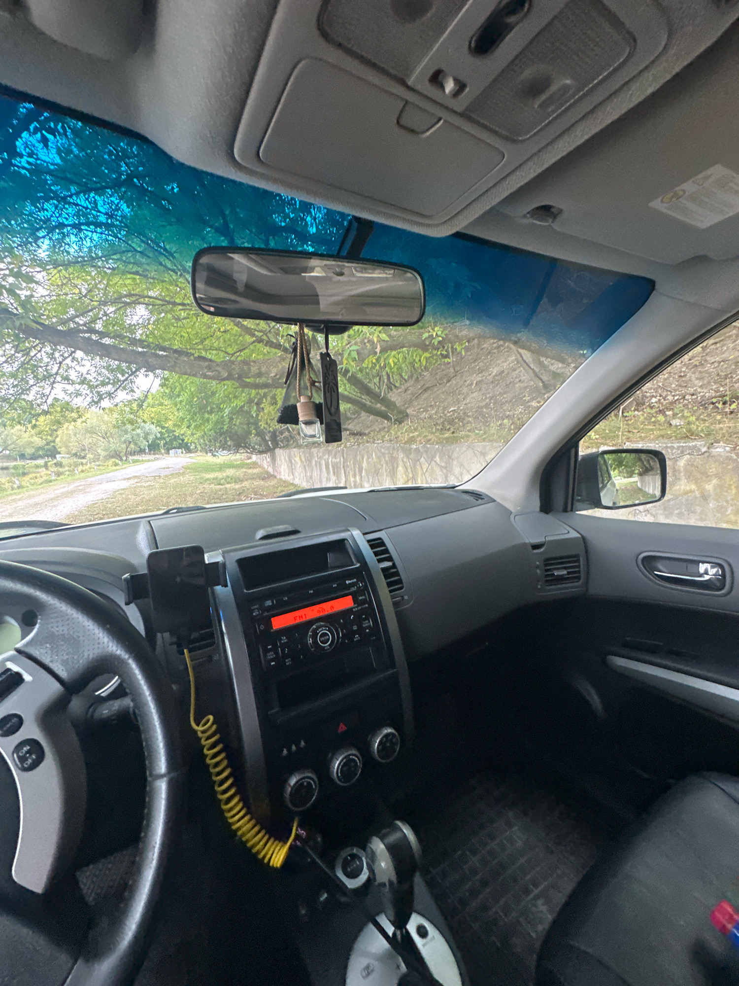 The image shows the interior of a car parked on a dirt road. The dashboard is visible, with a radio displaying "FM1 98.8". A phone is mounted on the dashboard. Outside the car, trees and a stone wall are visible. The scene appears to be in a rural area, possibly on the outskirts of Makiv, Ukraine, given the provided context. There are no people visible in the image. The car's interior is somewhat cluttered with air fresheners hanging from the rearview mirror and a coiled yellow cable. The lighting suggests it is daytime.