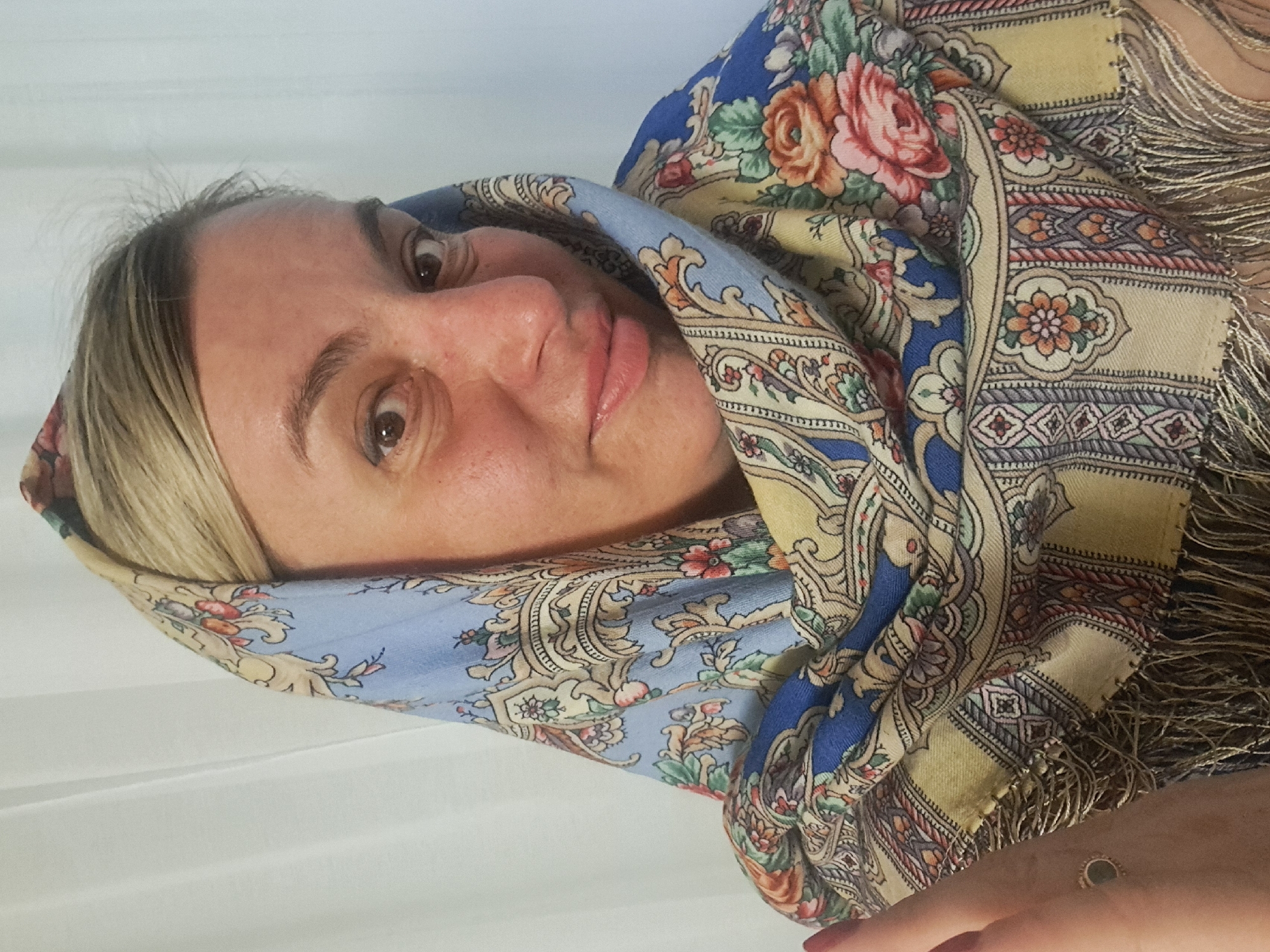 A person with fair skin and light blonde hair is wearing a patterned scarf or headscarf featuring floral and geometric designs in blue, yellow, and beige tones. The scarf frames the face and neck. The background appears to be a white, slightly wrinkled surface, possibly a curtain or fabric backdrop. The person is looking at the camera with a slight smile or smirk. No details suggest the city of Elektrostal, Russia.