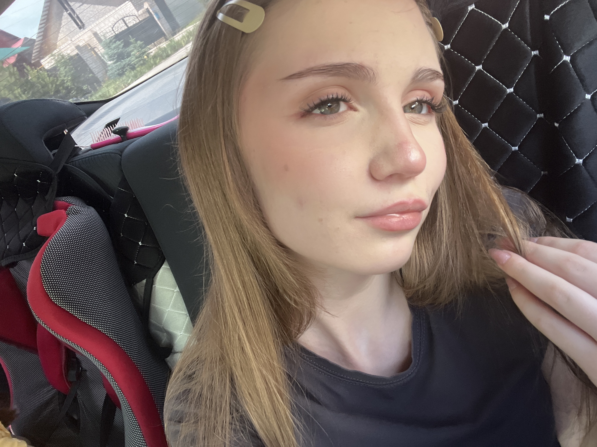 In an indoor setting, likely a vehicle, a young woman with long, light brown hair is captured in a close-up shot. She is looking towards the right, with her eyes open and her lips slightly parted. Her makeup includes blush on her cheeks and mascara on her lashes. A light-colored hair clip is visible on the left side of her head. The background reveals a dark, quilted fabric and part of a car seat with red and black accents. Outside the window, a building and trees are visible, suggesting an urban or suburban environment. The image does not provide any specific landmarks or details that would allow for identification of the city of Sterlitamak, Russia.