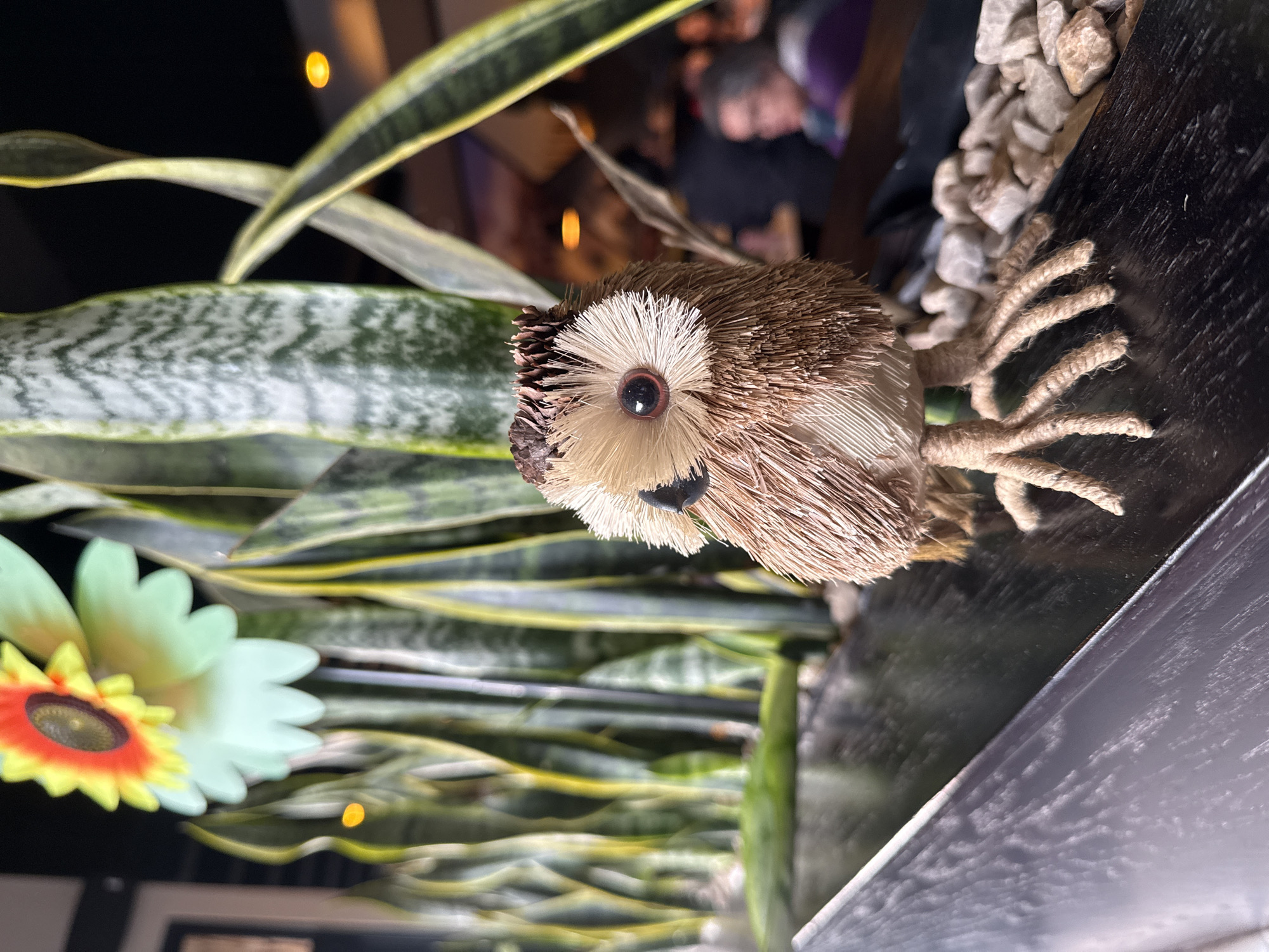 A straw owl figurine is in a display of potted plants, gravel, and a sunflower decoration in Mexico City, Mexico. The owl is brown and cream-colored with a black beak and a single visible eye. The legs are made of tan rope. The plants are snake plants. A blurred person is in the background.