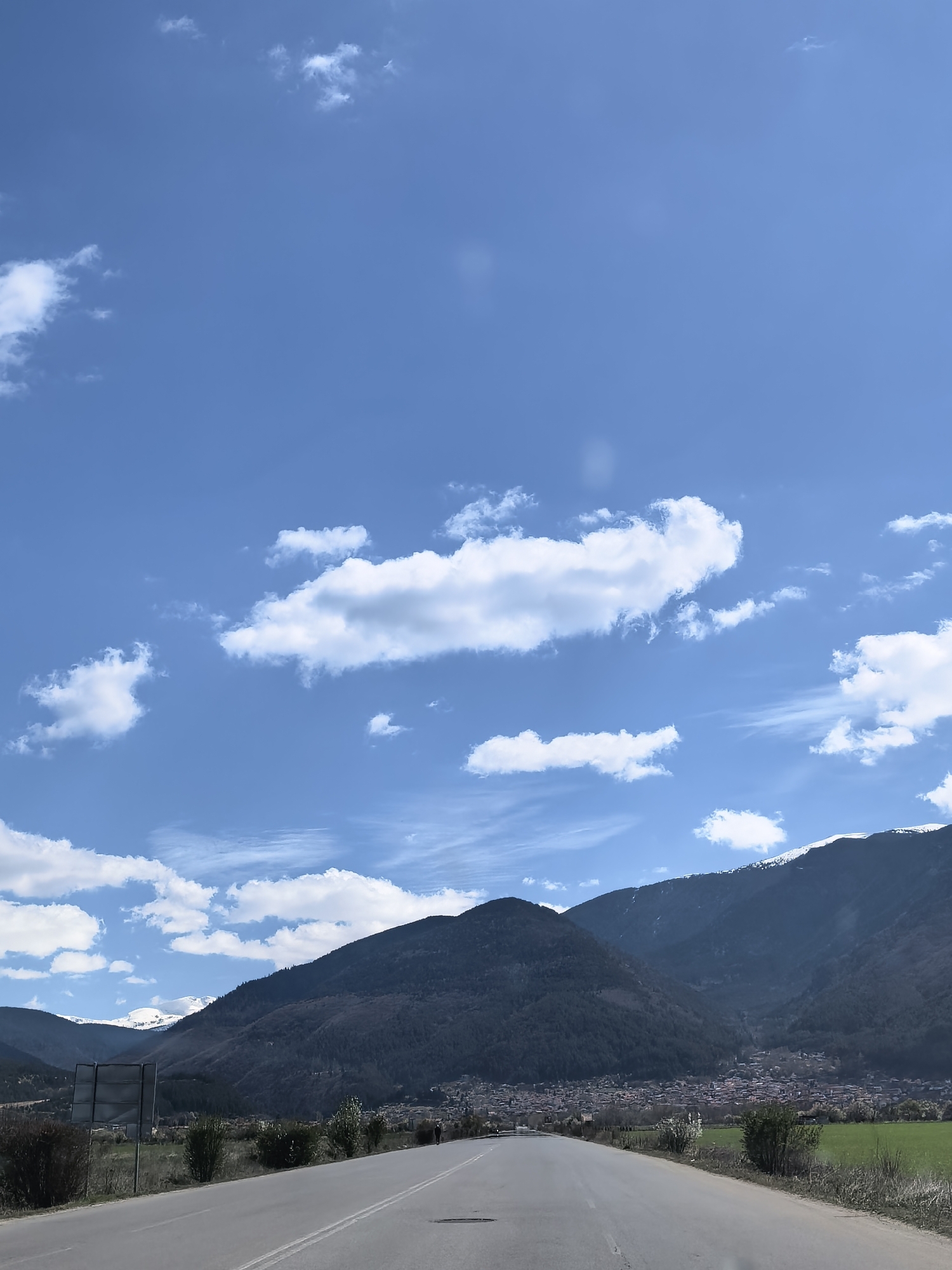 The image shows a road leading towards a town nestled in a valley between mountains. The sky is blue with scattered white clouds. The mountains are dark, with some visible bare trees and possibly some snow on the peaks. The road is gray, with white lines marking the lanes. There are green fields on either side of the road near the camera. The town appears to consist of many closely packed buildings with reddish roofs. There is a sign to the left of the road in the midground. The location is Unknown, Unknown.