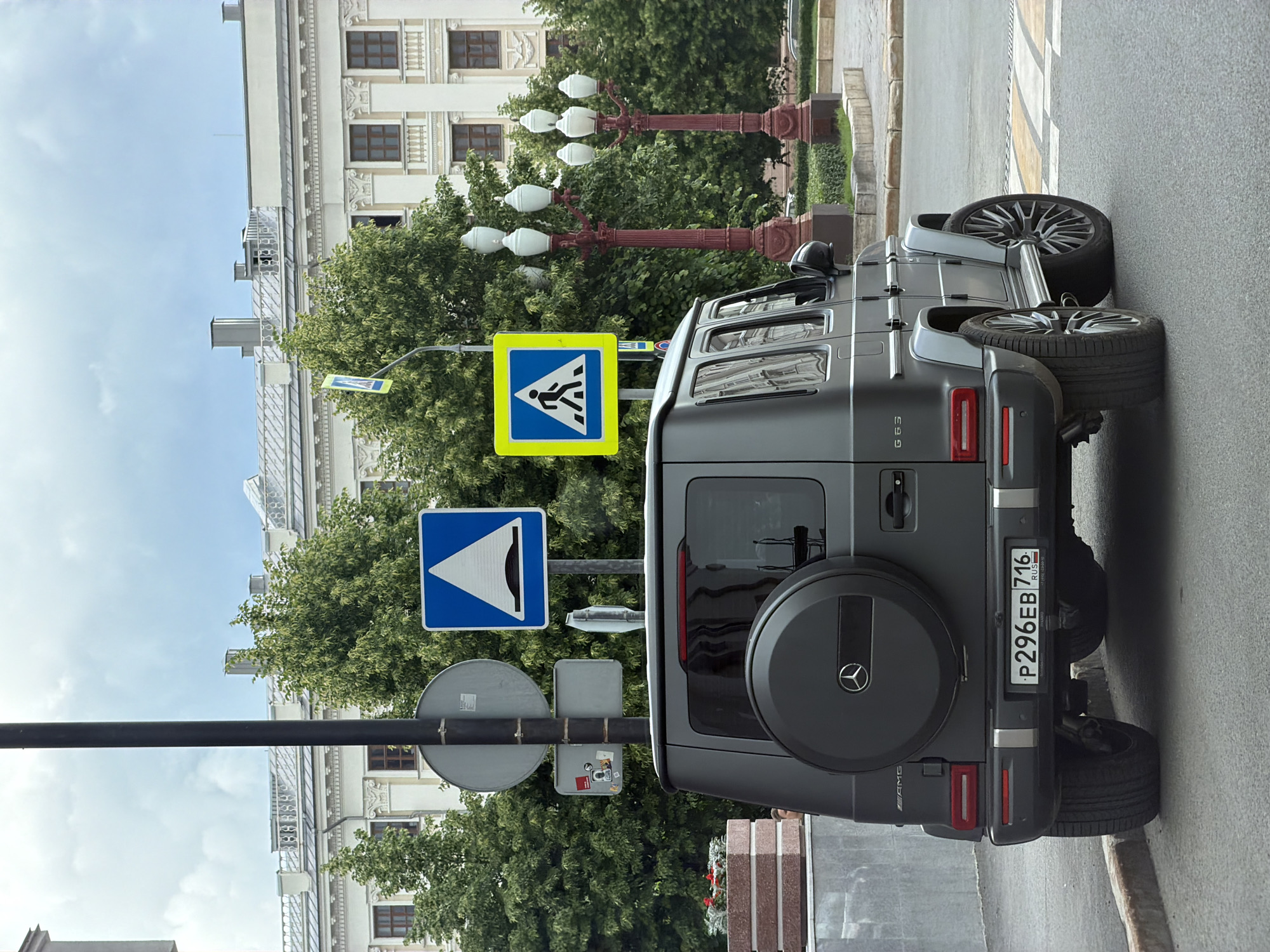 The image shows the rear of a dark gray Mercedes-Benz G-Class SUV parked on a street in Kazan, Russia. The vehicle's license plate reads "P296EB 716 RUS". Several traffic signs are visible in the background, including a pedestrian crossing sign, a yield sign, and a roundabout sign. A building with ornate architectural details is visible in the background, along with trees and lampposts. The sky is partly cloudy.