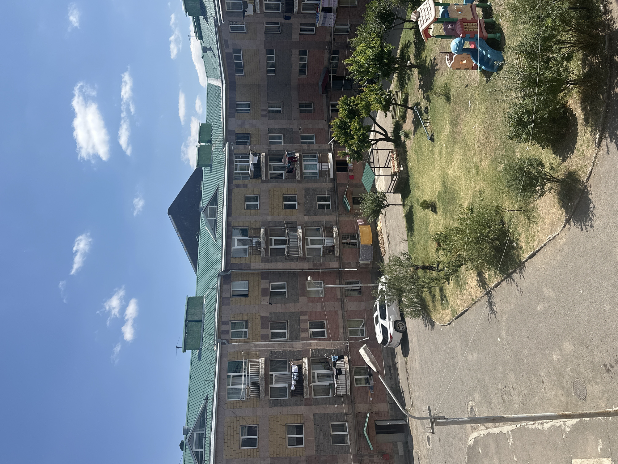 The image shows a courtyard of an apartment complex in Gyumri, Armenia. The complex is a multi-story building with a green metal roof. The facade of the building is constructed with a mix of brick and stone in various colors. Balconies with metal railings are visible, some of which have laundry hanging out to dry. A white SUV is parked on the asphalt courtyard. The courtyard also features a playground with a slide and other colorful structures, as well as trees and bushes. A street lamp stands in the foreground. The sky is blue with scattered white clouds.