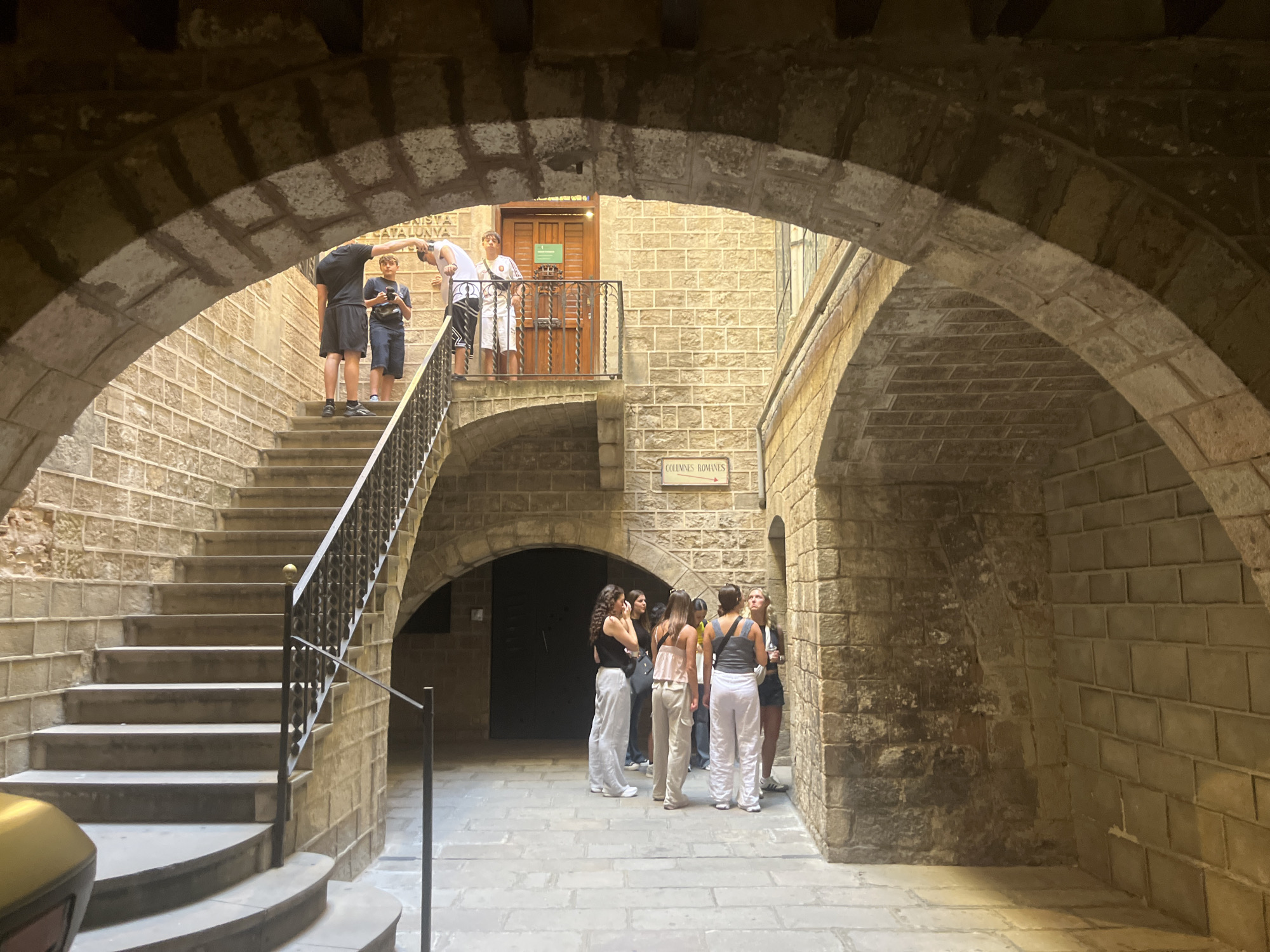 The image shows a stone courtyard in Barcelona, Spain. An arched stone entrance frames the scene. Several people are present. On the left, a group of boys and a man are standing on a stairway, while another group of people, primarily women, are standing on the courtyard ground. A sign reads "Col·lumnes Romanes" ("Roman Columns" in Catalan). The building's architecture is characterized by exposed stone, arches, and staircases.