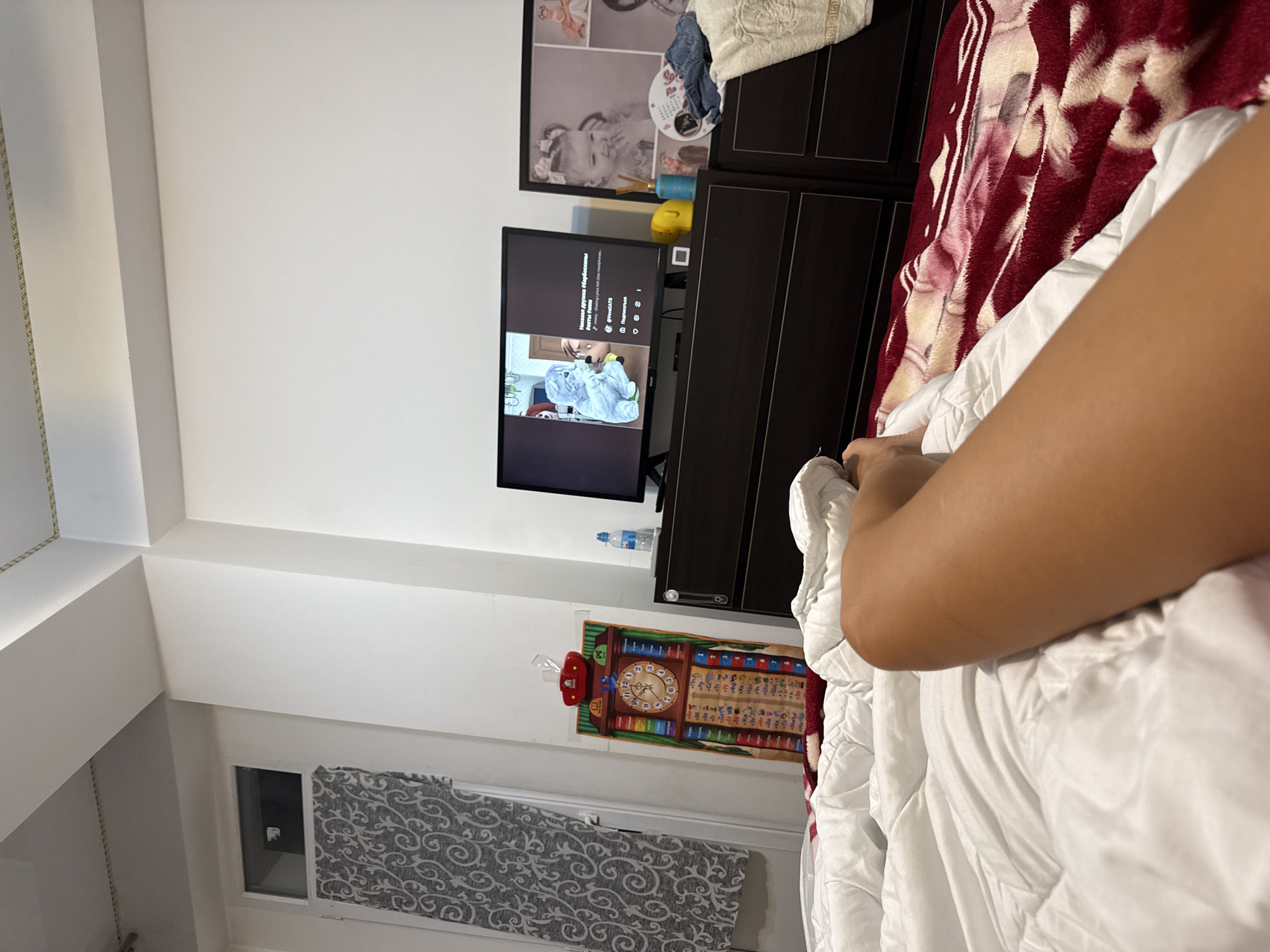 The image depicts an indoor scene, likely a bedroom, with a person's leg visible in the foreground, resting on a white duvet atop a red and white patterned blanket. A television screen displays a video with animated characters and Russian text. To the left of the television, a dark dresser holds a water bottle, a yellow toy, and a diffuser. To the left of the dresser, a colorful educational mat resembling a grandfather clock hangs on the wall. Further left, a door with a window covered by a patterned curtain is visible. The environment suggests a home setting, possibly in Chișinău, Moldova, given the presence of Russian text on the television and the style of the educational mat.