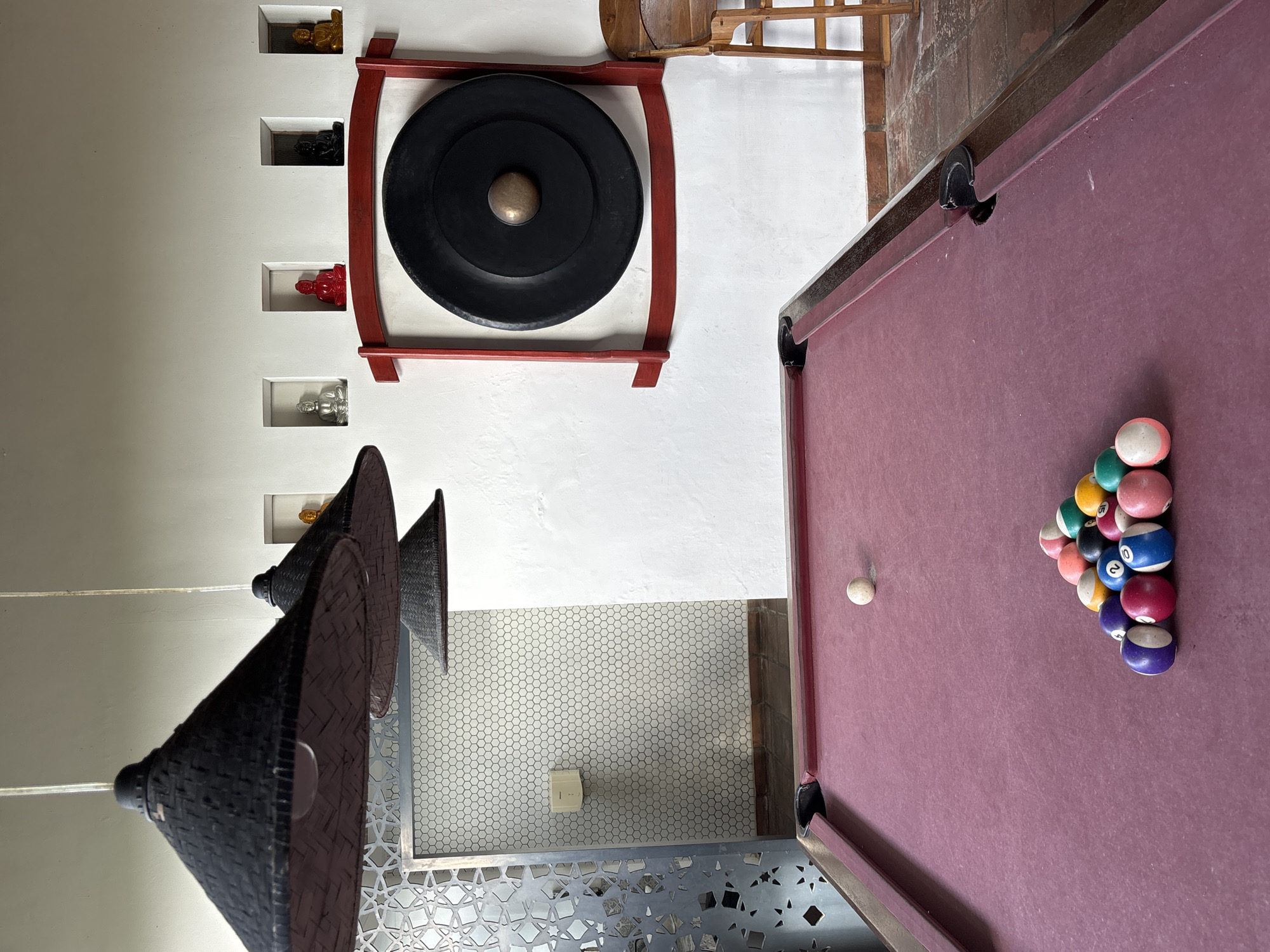 The image depicts an indoor scene in Kuta Selatan, Indonesia, featuring a pool table with a racked set of pool balls and a cue ball on a red felt surface. Above the pool table are three conical lamps with a woven texture. A large gong in a red frame is mounted on a white wall alongside recessed niches holding small figurines. The floor near the pool table is tiled in a small hexagonal pattern and features decorative metalwork.