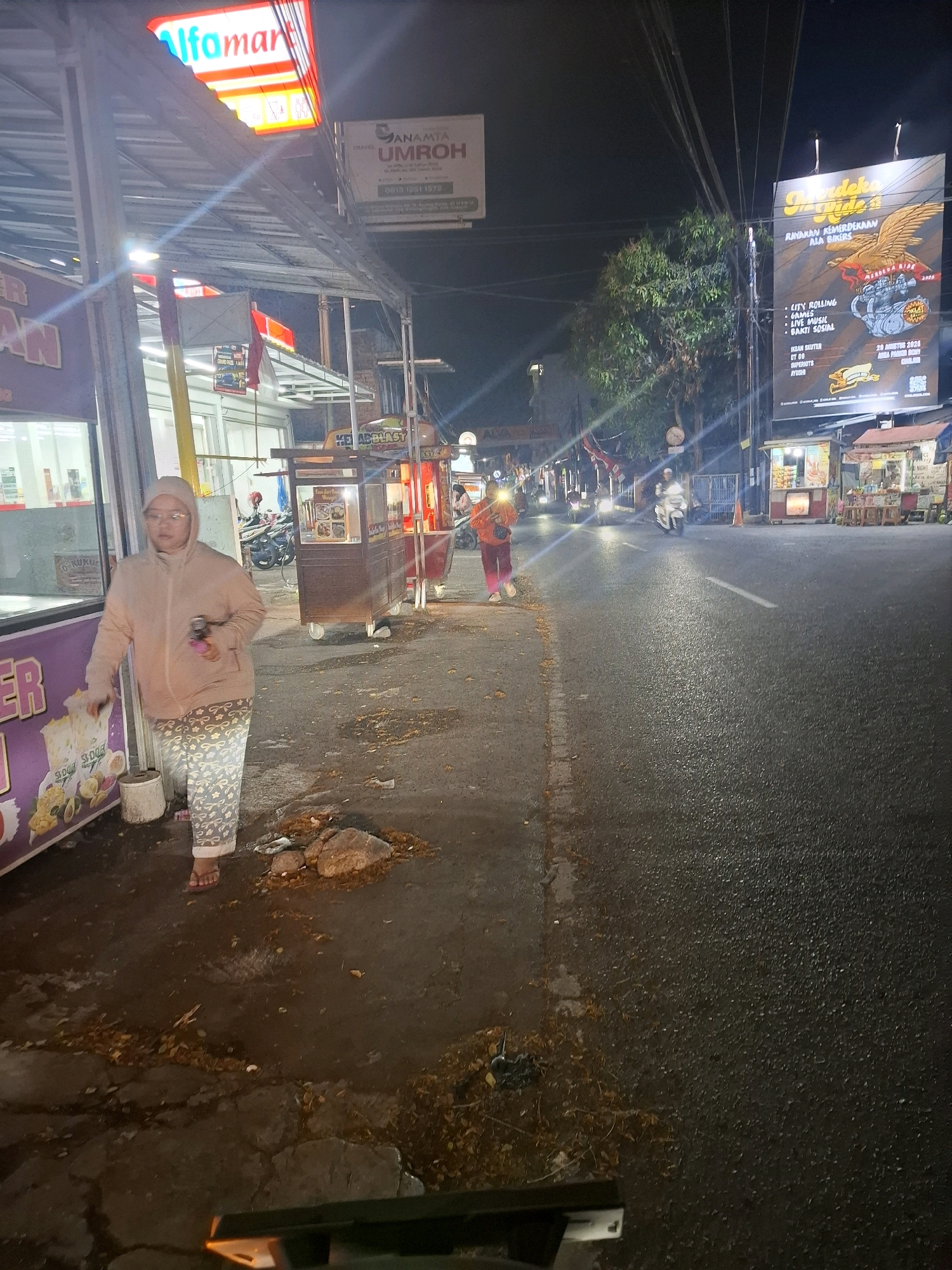 The image depicts a street scene at night in Bojong, Indonesia. On the left, a person wearing a light-colored hooded sweatshirt and patterned pants walks on the sidewalk. They are carrying a pink object in their right hand. Adjacent to the sidewalk is a storefront with an "alfamart" sign illuminated in red and white. To the right of the person, a food stall with a brown wooden structure is visible, with its wares displayed under lights. Further down the street, other food stalls and shops are open, with some patrons milling about. Vehicles, including motorcycles, are present on the road. A large billboard with an eagle graphic and text in Indonesian is displayed on the right side of the street. The road surface appears wet, suggesting recent rain or a damp environment. Fallen leaves are scattered on the sidewalk and road. The overall atmosphere is that of a bustling urban street at night.