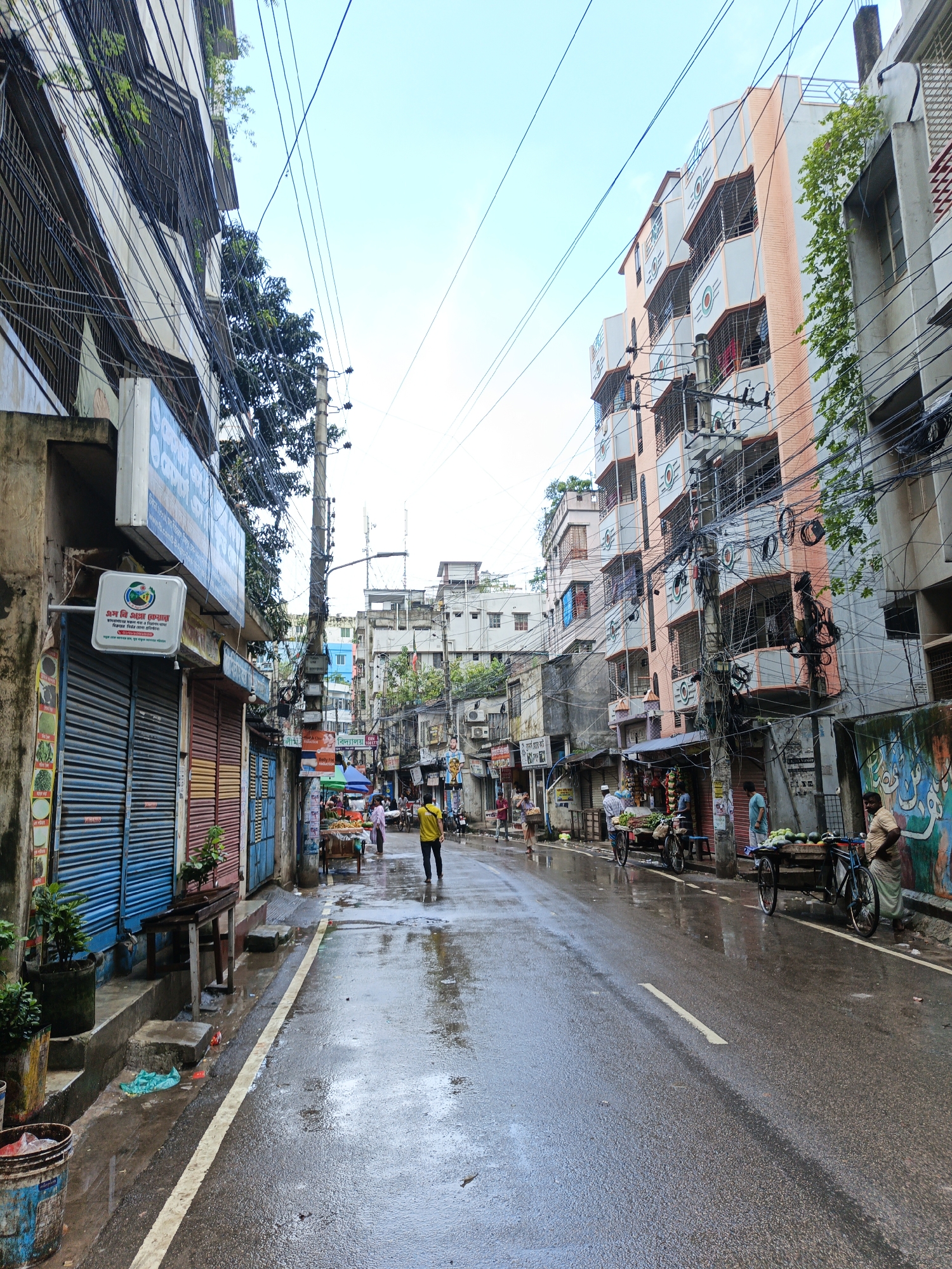 A street in Dhaka, Bangladesh, is wet and reflects the sky. Buildings line both sides of the street, with a mix of shops on the ground floor and apartments above. Overhead, a tangled mess of electrical wires crisscrosses the sky. On the left, a building has a blue shuttered shop and a sign with text in Bengali. On the right, a brightly colored building with balconies stands tall. Several people are present. A man in a yellow shirt walks down the middle of the wet street. Further down the road, vendors are selling produce from carts. One cart is laden with watermelons. Another cart is visible behind it, piled high with more fruits and vegetables. A woman in a sari stands next to a cart. The overall scene depicts a typical bustling street in Dhaka after a rain shower.