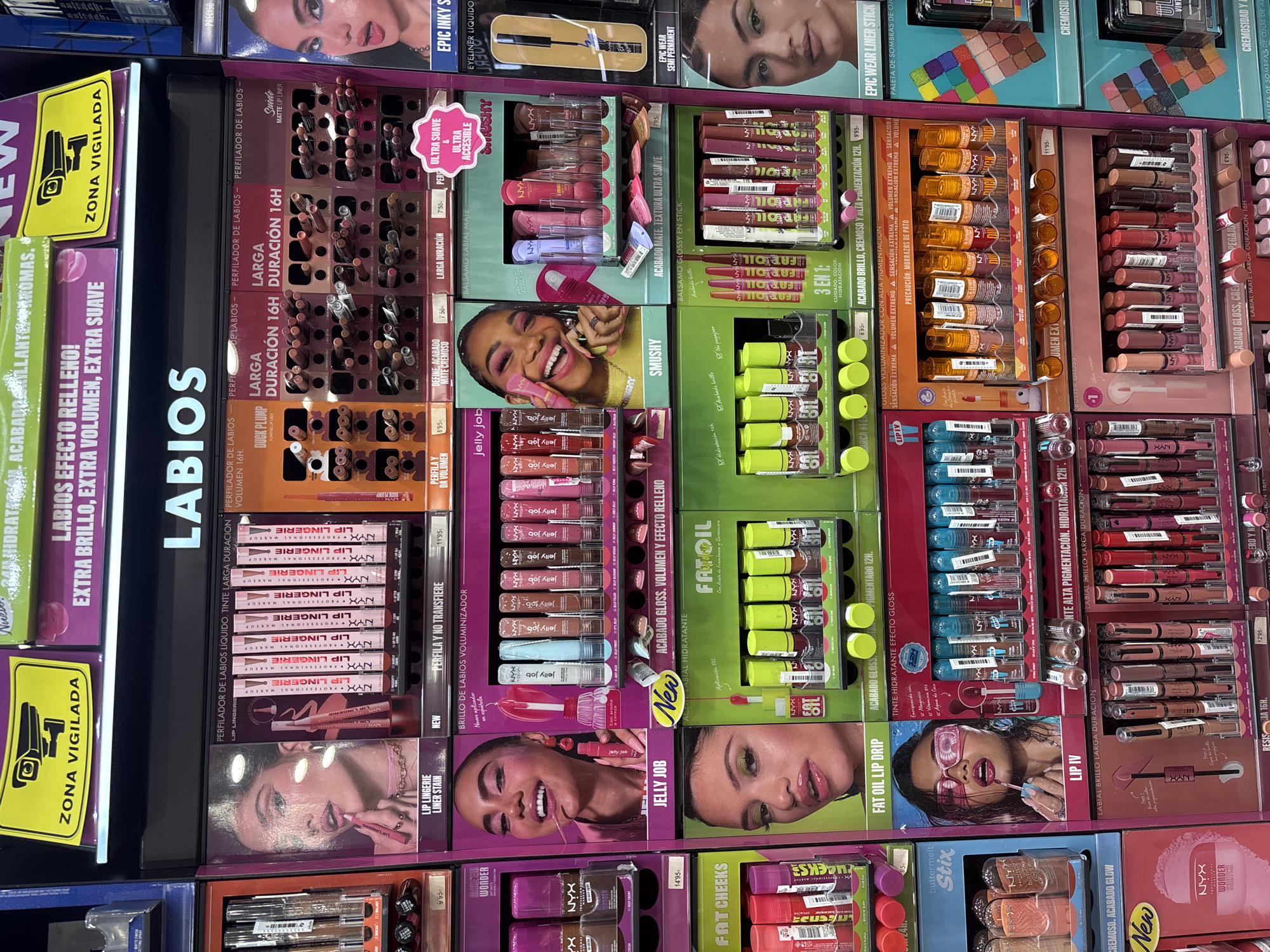 The image depicts a brightly lit, indoor retail display of makeup products, primarily lip products from the brand NYX Professional Makeup. The display is organized in a tiered, wall-mounted format, with various sections showcasing different product lines. Prominent signage in Spanish indicates product features like "LARGA DURACIÓN" (long-lasting), "EFECTO RELLENO" (plumping effect), and "EXTRA BRILLO" (extra shine). Several product categories are visible, including lip liners, liquid lipsticks, lip glosses, and lip stains, distinguished by their packaging and product names. The background is a mix of purple, green, and pink signage and product displays. The overall impression is that of a cosmetics section within a larger store or a dedicated brand display. Without additional information or context specific to Orihuela, Spain, it is not possible to definitively locate this scene within that city. The image focuses solely on the retail display and its contents.