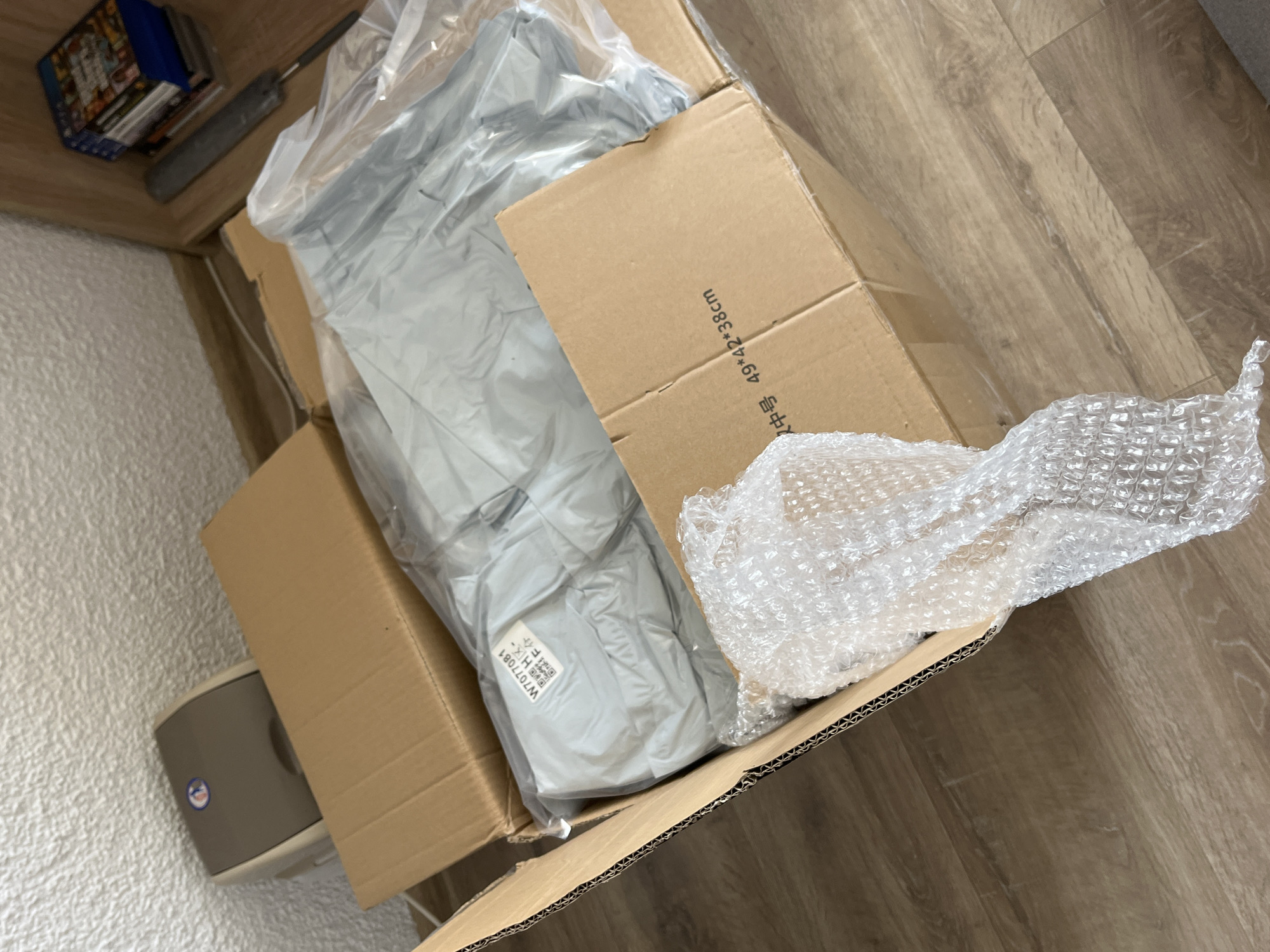 The image shows an open cardboard box sitting on a light brown wooden floor. Inside the box are various items, including an item wrapped in gray plastic, bubble wrap, and a smaller cardboard box with Chinese writing "尺寸 49x42x38cm" on the side. In the upper left corner of the image, a stack of game cases and a gray cylindrical object are visible on a light-colored shelf or table. To the left of the box there is a gray air freshener. The location in the city of Zlatograd, Bulgaria, is not discernible from the image.