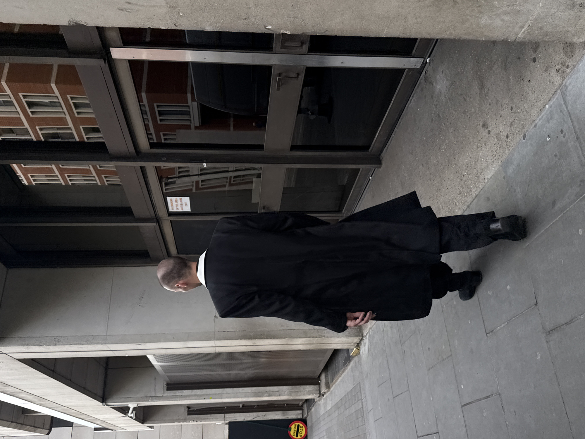A man in a black suit and coat walks past a building. The building has a glass door reflecting the brick building opposite. The location is London, United Kingdom, as evidenced by the "Stop Works" sign on the ground.