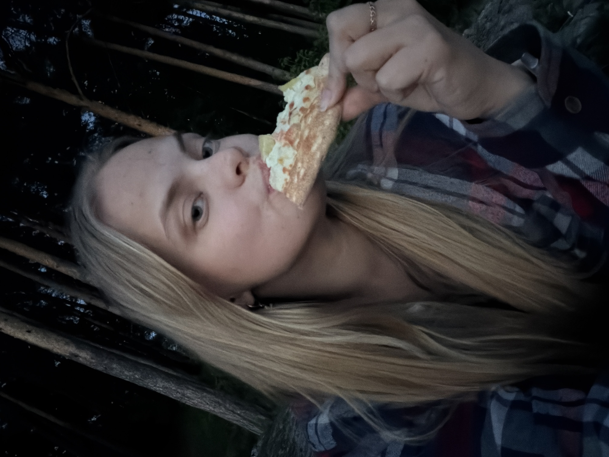 A person with blonde hair, wearing a plaid shirt, is holding and eating a piece of pizza. The background is dark with glimpses of wood, potentially in a wooded area. There is no indication of Izhevsk, Russia.