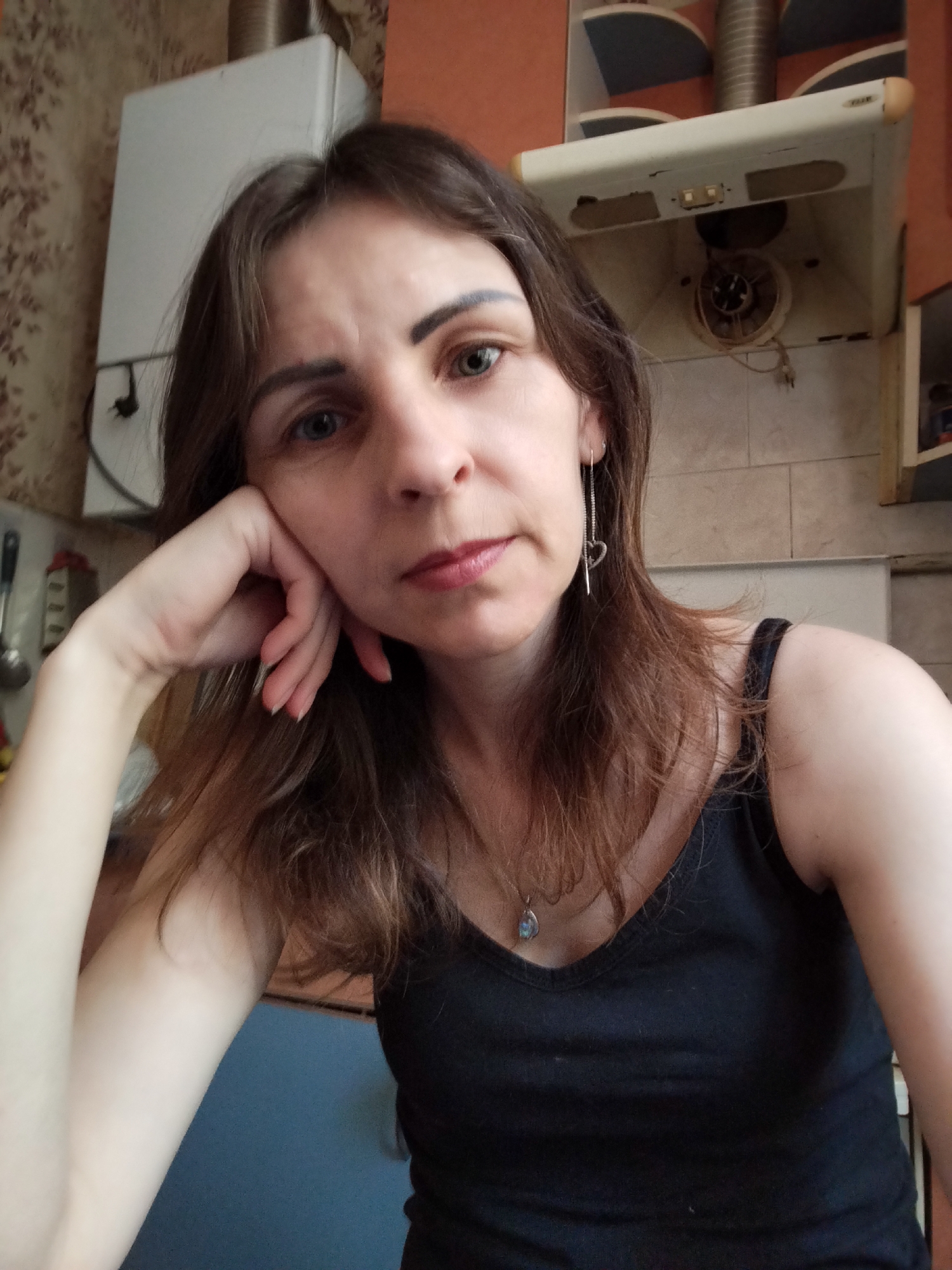 A medium shot captures a woman with shoulder-length brown hair and blue eyes, resting her head on her left hand. She is wearing a black tank top and a silver pendant necklace with a blue stone, as well as a long silver earring with a heart charm in her right ear. The background appears to be a domestic kitchen with a white gas water heater on the left and a beige range hood above a stovetop on the right. The woman's expression is neutral. The image does not provide any specific information about the location within Vinnytsia, Ukraine.