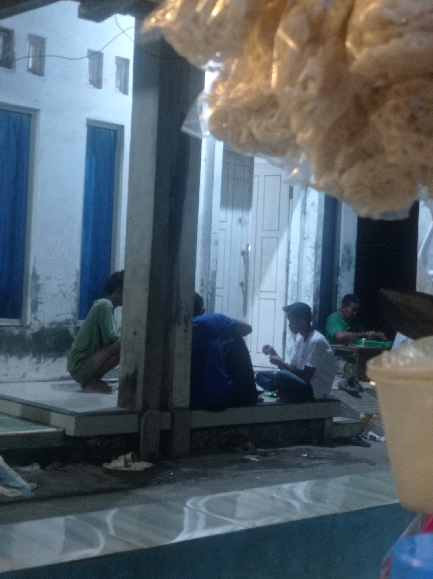 The image shows a group of people near a building with blue curtains. In the foreground, there are several individuals sitting on a low ledge. One person is wearing a green shirt and shorts and is in a squatting position. Another person is wearing a blue shirt and appears to be sitting. A third person is wearing a white t-shirt and jeans and is looking down. In the background, a person in a green shirt is seated at a small table, and there is another person visible behind them. The environment appears to be outdoors, with a concrete floor and some debris on the ground. The building has several windows and a door. The overall lighting suggests it is evening or a dimly lit area. The image quality is low, with some blurriness and graininess. This scene is likely taking place in a residential area or an outdoor communal space in Babalan, Indonesia.
