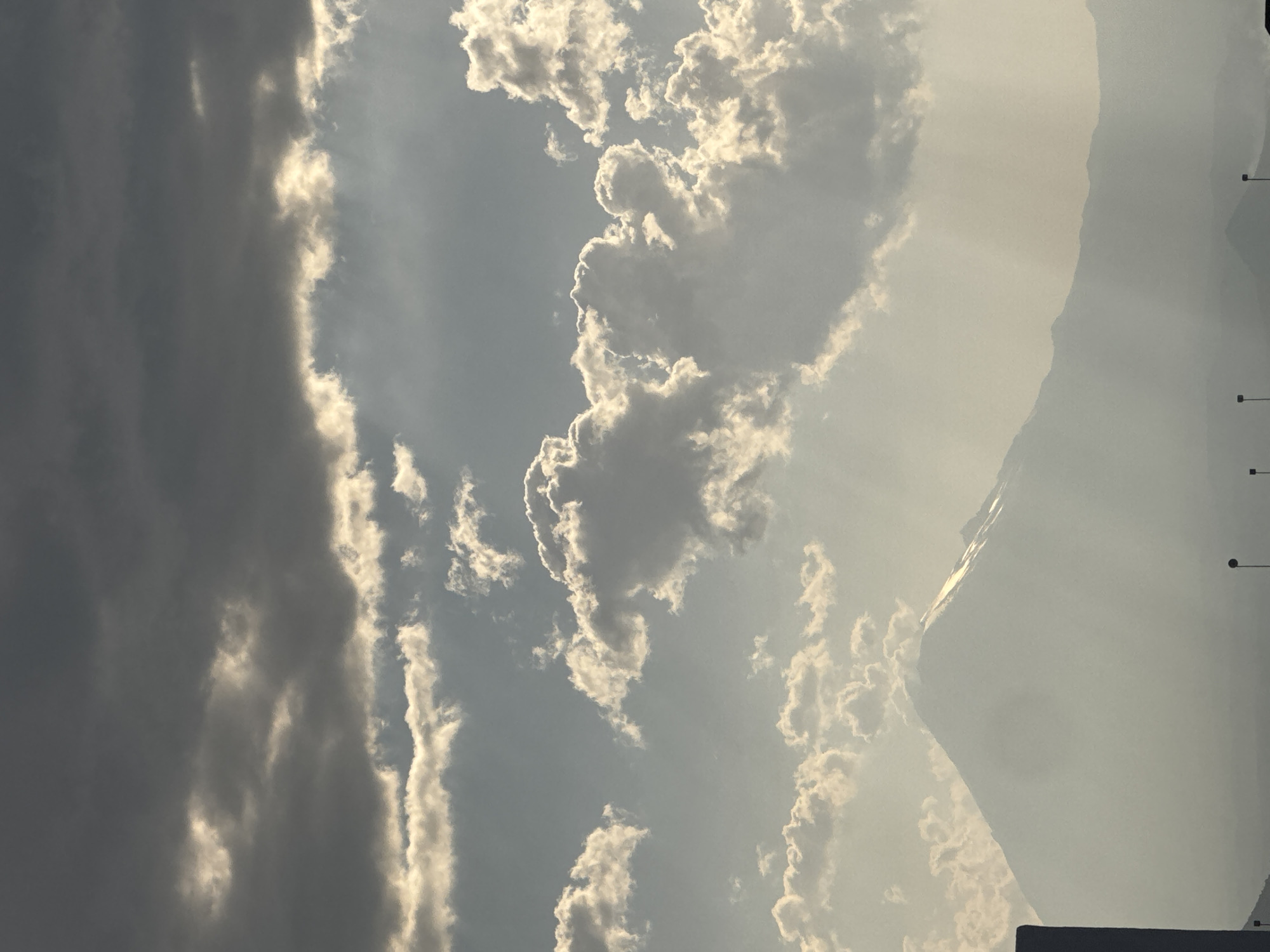The image depicts a panoramic view of a cloudy sky and mountainous terrain, likely captured from San Francisco Ocotlán (Ocotlán), Mexico. The upper third of the frame is dominated by dark, dense stratocumulus clouds, transitioning to brighter, more broken cumulus clouds in the middle ground, where sunlight is prominently breaking through, creating visible light shafts. In the mid-distance, a large, snow-capped mountain or volcano rises prominently, its peak illuminated by the sunlight, appearing hazy against the bright sky. The lower foreground features the silhouetted top of a dark, vertical architectural structure on the far left, and a series of at least four dark, pole-like objects, consistent with streetlights or utility poles, extending across the bottom of the frame. The overall atmosphere is one of dynamic natural light under variable cloud cover, highlighting the mountain landscape. No people are visible.