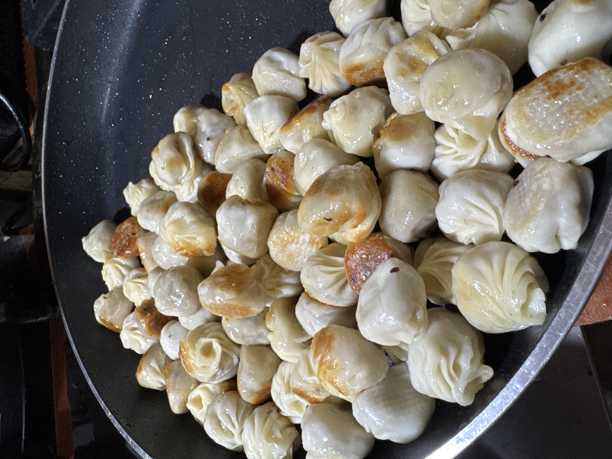 The image shows a large pan filled with numerous steamed dumplings. Some dumplings have a golden-brown, slightly crispy bottom, suggesting they were pan-fried or seared briefly after steaming. The dumplings are mostly round and slightly wrinkled in appearance, with some showing a visible swirl pattern from how they were folded. They appear moist and possibly glistening from steam or a light sauce. The pan is dark, likely non-stick, and the dumplings are densely packed within it. The image gives no information to associate it with the Wulong District of China.