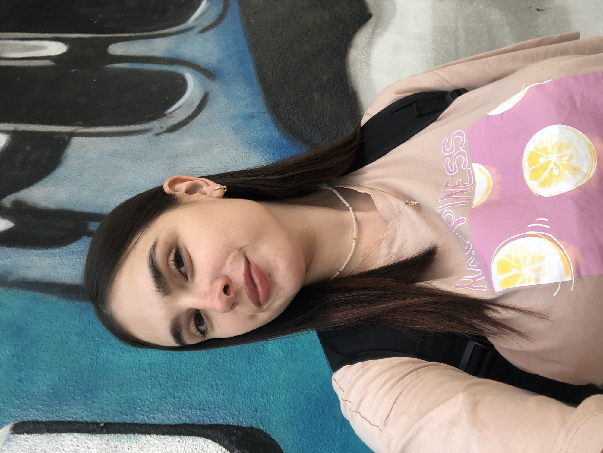 A young woman with long, dark hair is taking a selfie outdoors against a graffiti-covered wall. She is wearing a light pink t-shirt with a graphic design featuring lemons and the word "HAPPINESS" in pink lettering. She also has a pearl necklace and a small gold pendant. The wall behind her is predominantly blue with black and white graffiti. The image does not contain any information that would indicate the location in the city of Chaikovsky, Russia.