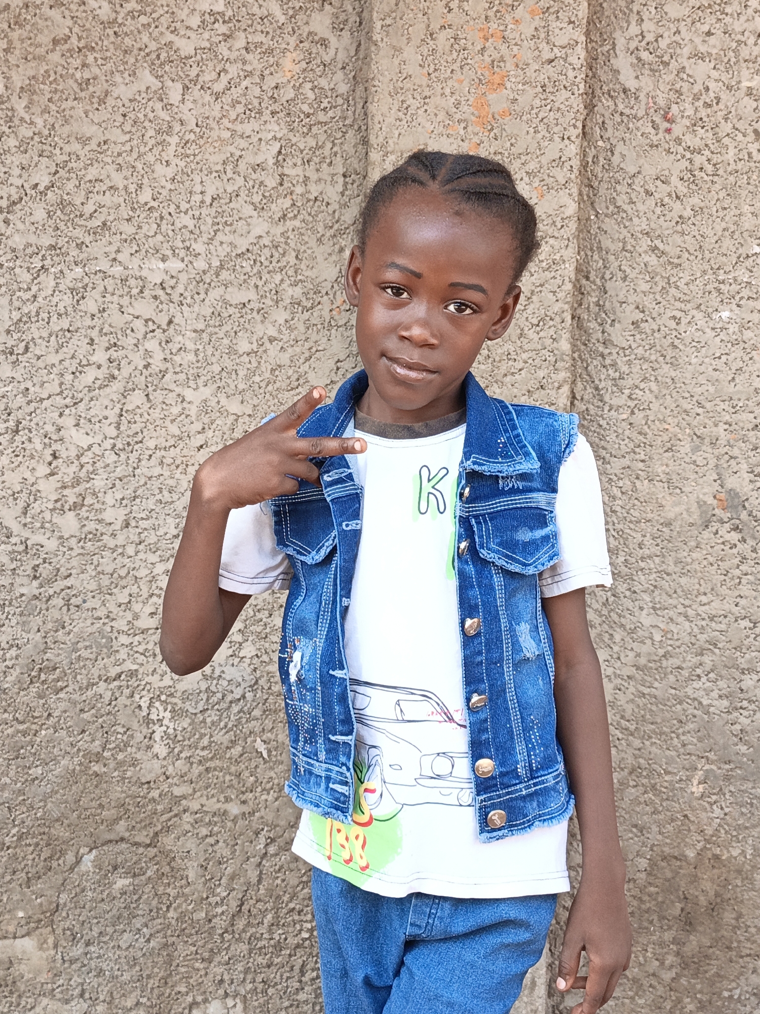 A young Black girl stands facing the camera in front of a rough, light-colored wall. She wears a white t-shirt with a car graphic and a letter "K" on the front, a blue denim vest with frayed edges and silver buttons, and blue jeans. Her hair is braided and styled away from her face. She is making a peace sign with her left hand. The location is Gwagwarwa, Nigeria.