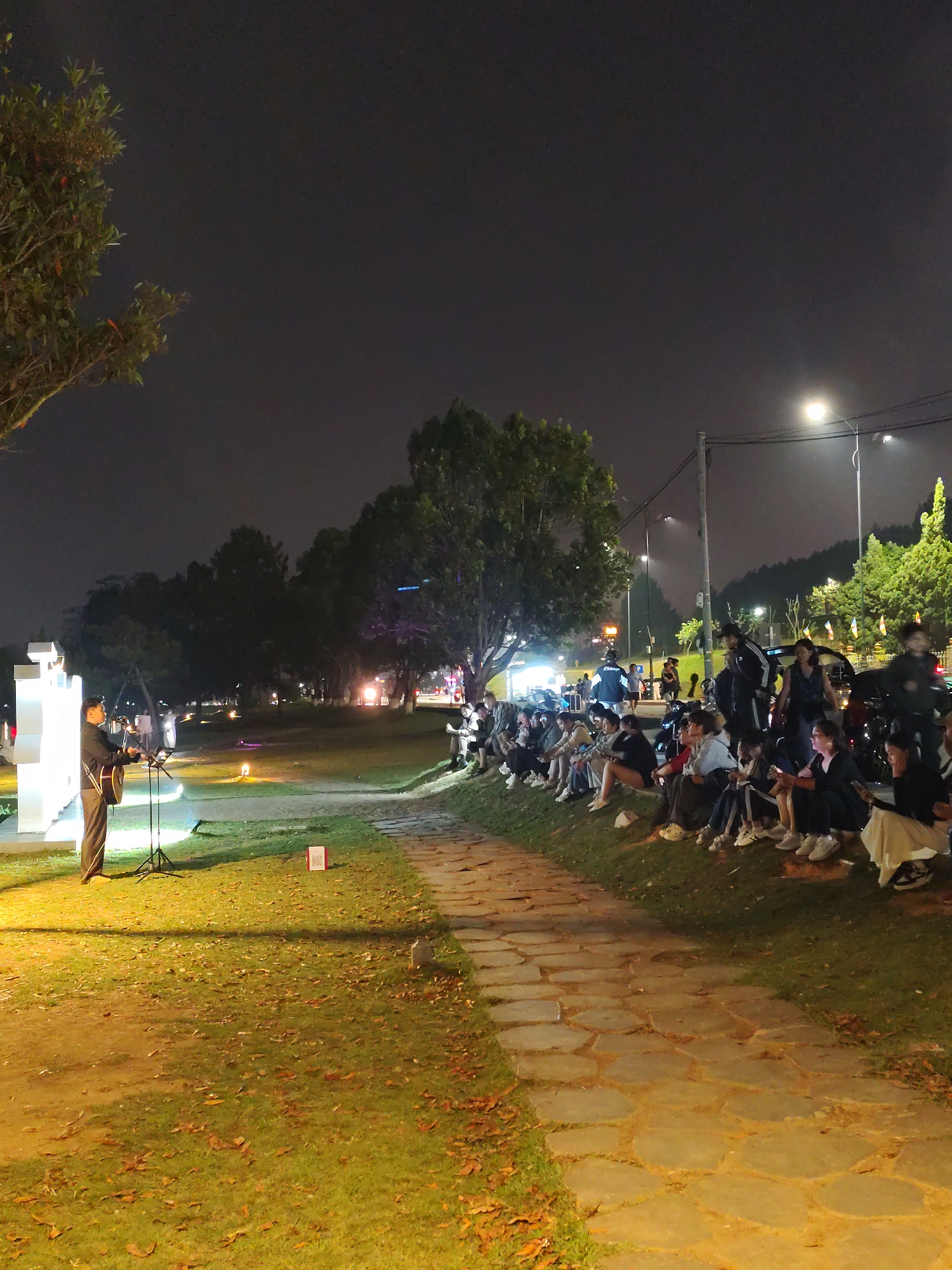 The photograph shows an outdoor scene at night. A crowd of people, predominantly young adults, sit on a grassy slope, seemingly watching a performance. A man stands near a brightly illuminated white structure, possibly art or architecture, and is playing a guitar. He has a microphone stand set up and appears to be performing. The area is lit by a mix of stage lighting, streetlights, and ambient light from the surroundings. A stone pathway leads toward the crowd. Trees and other vegetation are visible in the background. Based on the image content, the location is the city of Da Lat, Vietnam.