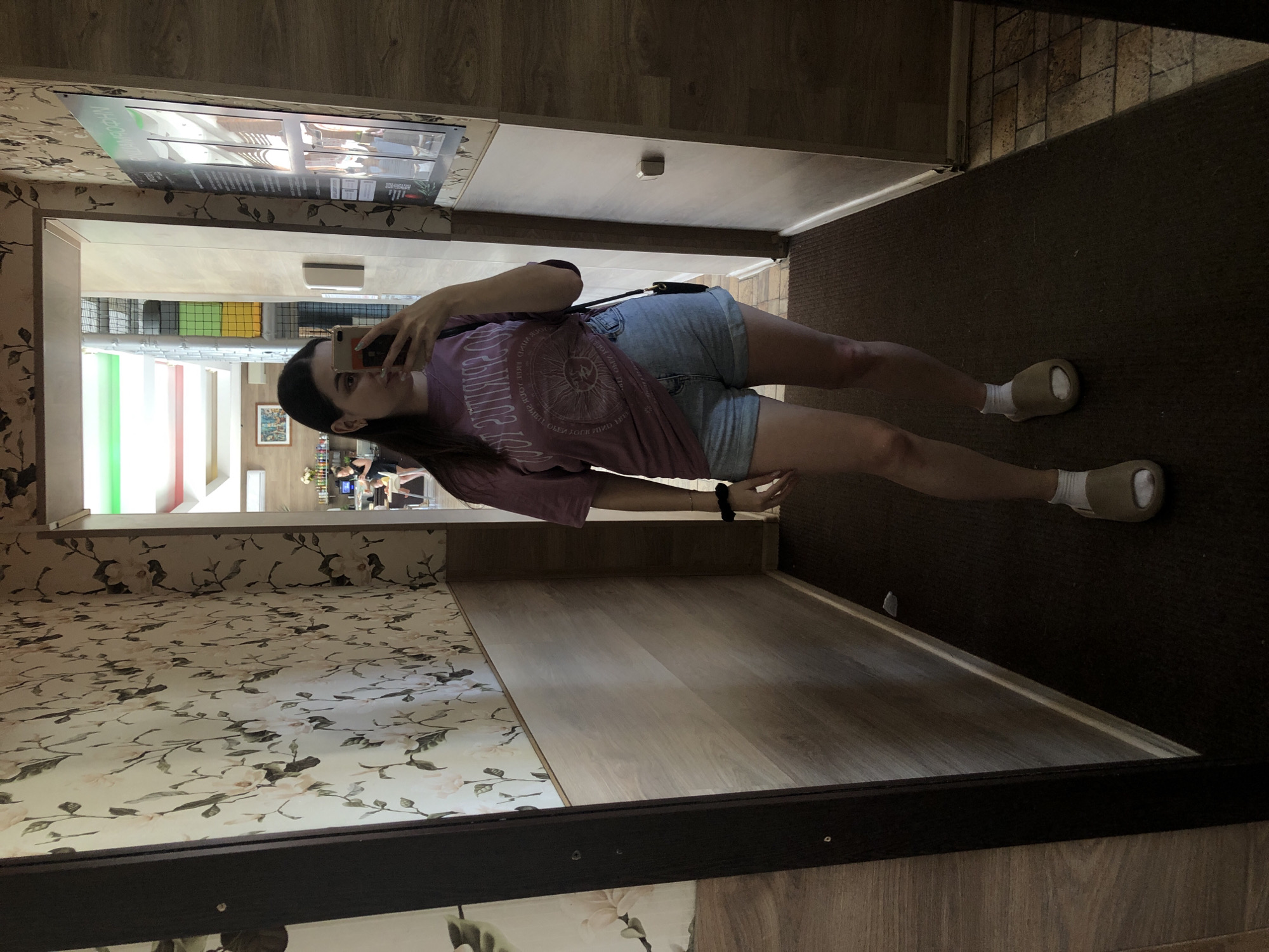 A woman is taking a selfie in what appears to be a hallway. The hallway has wallpaper on the left side with a floral pattern, while the right side leads to a brown-carpeted area. A poster or advertisement is on the wall above the hallway entrance. In the background, there's a glimpse of what looks like an interior space with colorful elements. According to the available information, the location is in the city of Chaikovsky, Russia. The woman is wearing a pink t-shirt and denim shorts with slide-on sandals and white socks. She has dark hair. She is holding her phone up to take the photo.