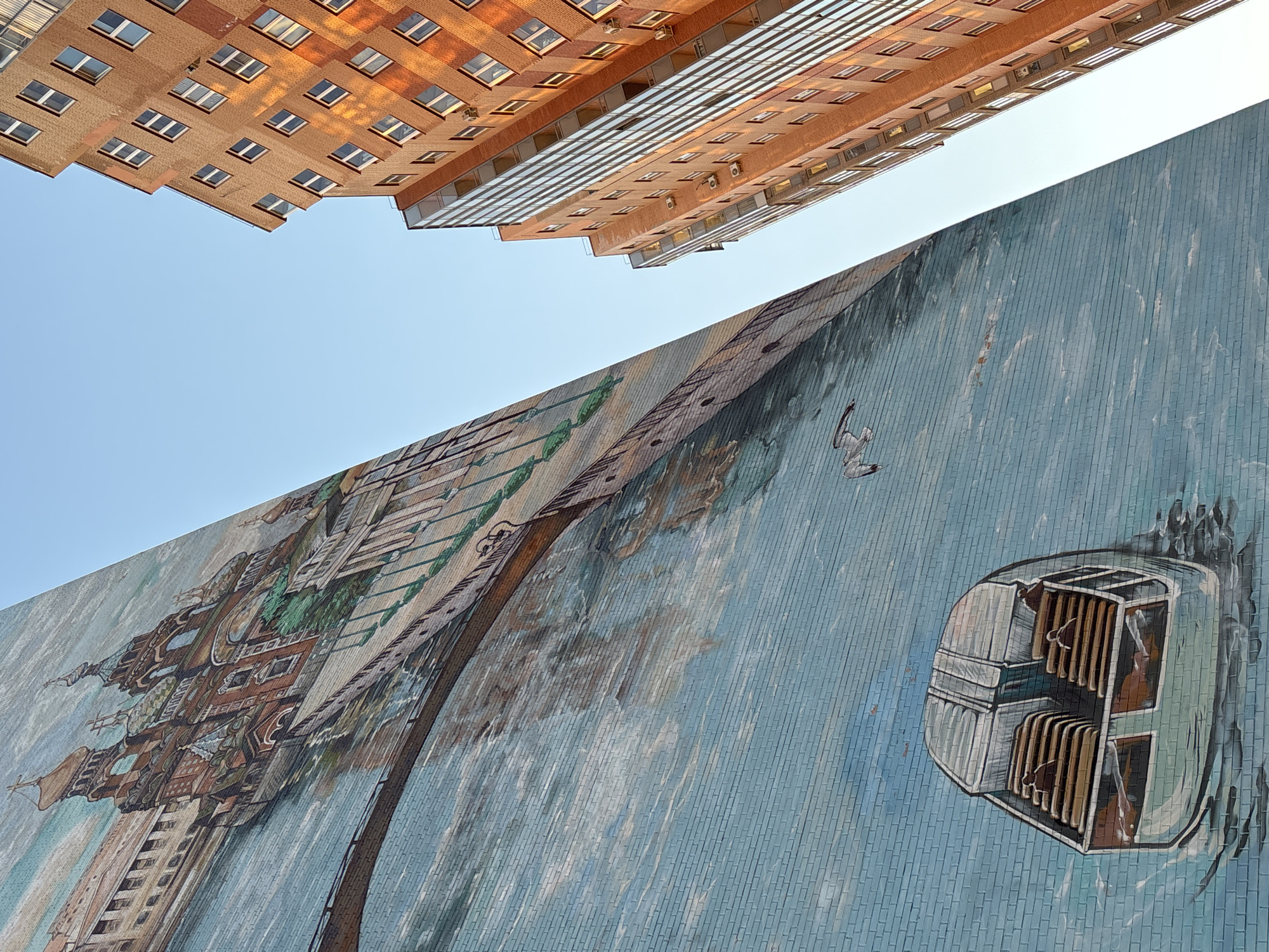 The image shows a low-angle view of two buildings in Kudrovo, Russia. The building on the left is an orange brick apartment complex with multiple windows. To the right is another building with a large mural painted on its side. The mural depicts a surreal scene featuring a detailed architectural structure resembling a castle and a water-like background. A futuristic-looking capsule or vehicle is also painted on the mural. Above the buildings is a clear blue sky.