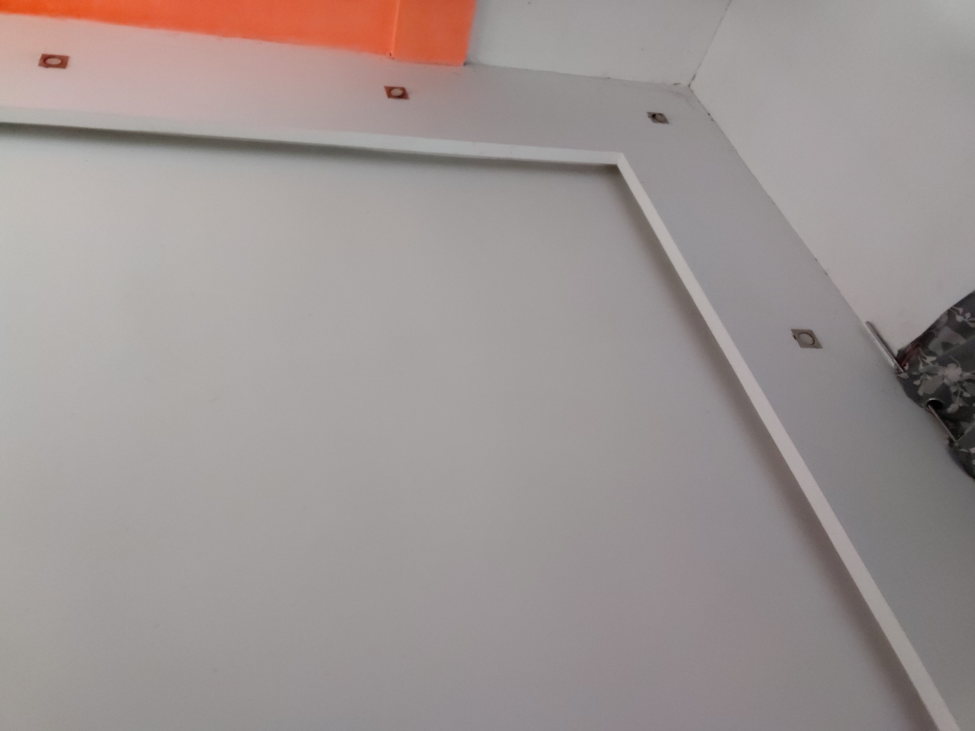 The image captures an upward-looking view of an indoor architectural space, likely a room or hallway, characterized by clean lines and neutral tones. The dominant feature is a large, light grey wall surface bordered by a white, raised frame-like structure. Above this frame, the ceiling is predominantly white, with a striking orange-painted section visible in the upper left corner.
Several small, square-shaped fixtures, which appear to be either recessed lighting units or their covers, are embedded in the white ceiling and upper wall area, suggesting a planned lighting arrangement. These fixtures are symmetrically placed along the architectural lines. To the far right, a small section of a patterned fabric, possibly part of a curtain, garment, or bag, with a grey, white, and black floral or camouflage design, is visible against a lighter wall.
No people are present, and no specific activity is taking place. The scene conveys a sense of stillness within a modern interior environment, possibly located in Santa Ana, Venezuela. There is no visible text in the image.