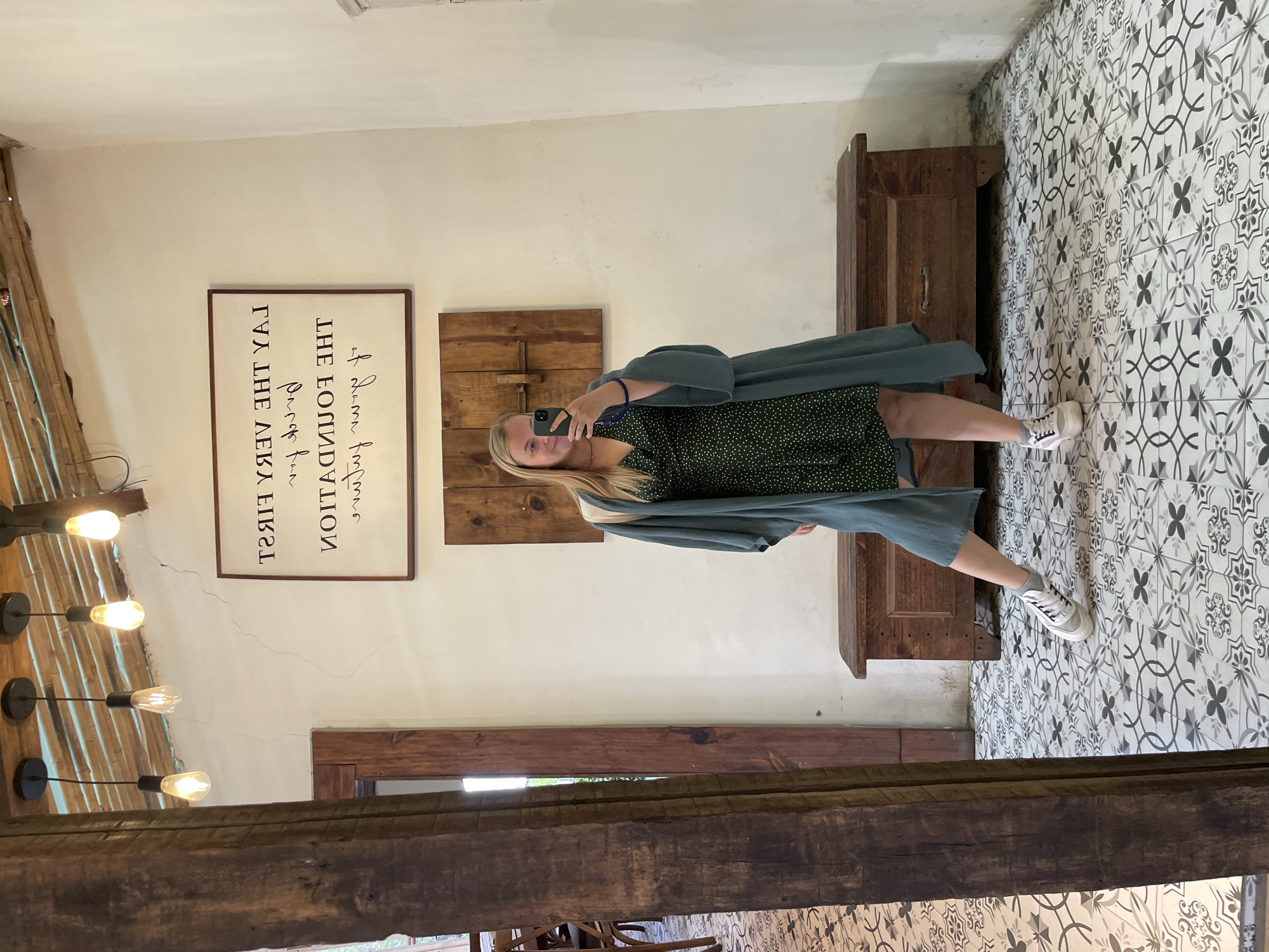A young woman with blonde hair, wearing a dark green polka dot dress and a teal kimono-style robe, stands in a room and takes a mirror selfie with her phone. She is wearing white sneakers and grey socks, and has a blue bracelet on her left wrist. The room has a white wall, a tiled floor with a black and white pattern, and a wooden bench against the wall. A framed quote hangs on the wall. The image appears to be taken indoors, possibly in a cafe or shop. It is not possible to determine the specific city or location within Phường Xuân Hương - Đà Lạt, Vietnam from the image alone.