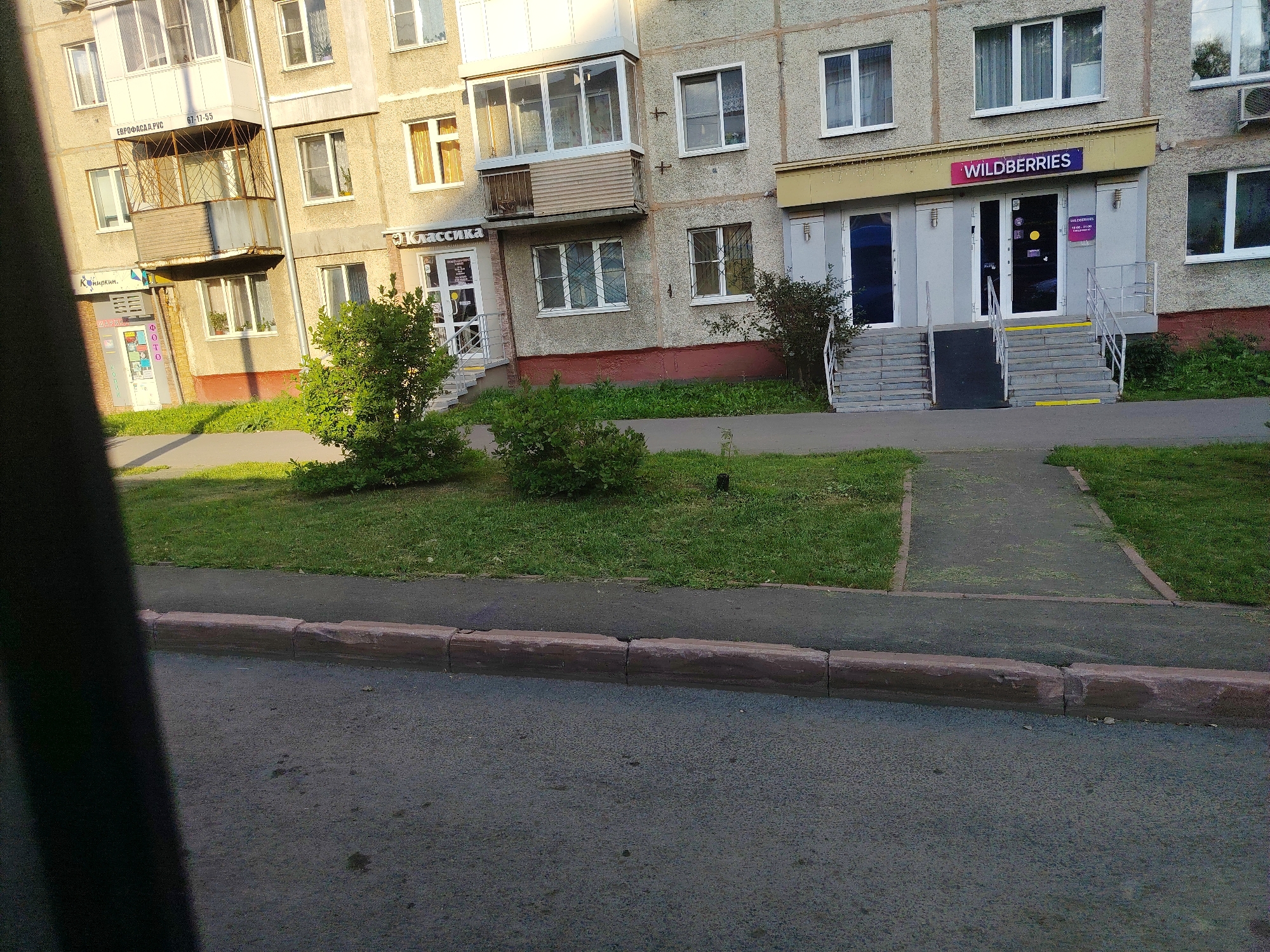 The image shows the exterior of a residential building with commercial establishments on the ground floor in Kemerovo, Russia. On the left, there is a store with a sign that appears to read "Капитолий" and another sign with Cyrillic text. Further to the right, there is a store with a sign that says "Классика". To the right of that, a store with the prominent "WILDBERRIES" logo is visible. The building has multiple stories with various windows and balconies. In front of the building, there is a sidewalk and a patch of green grass with some bushes and trees. The ground in front of the sidewalk is paved, likely a road or parking area. The overall scene depicts a typical urban environment with a mix of residential and retail spaces.