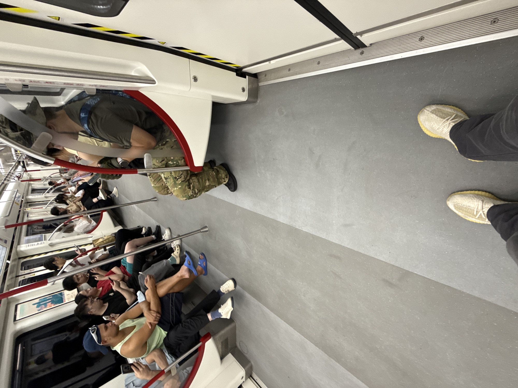 The image depicts the interior of a subway train in Baiyun District, China. Several passengers are seated on benches, engaged with their mobile phones or conversing. The train carriage features a neutral color palette with grey flooring and white walls, accented by red poles. One passenger in the foreground is wearing camouflage pants and a hat. Another passenger in the foreground is wearing a light green tank top and dark shorts, with blue flip-flops. The floor is a light grey, and the lighting appears to be artificial.