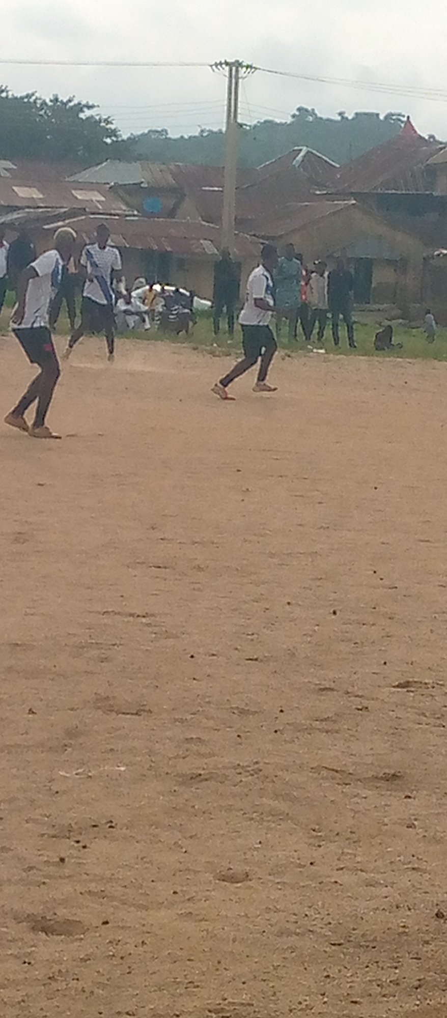An outdoor scene captures a group of individuals on a dirt field, with buildings and trees in the background under a cloudy sky. The foreground is dominated by the rough texture of the dirt ground, with subtle variations in color and scattered small pebbles. In the middle ground, two young males in athletic attire are engaged in what appears to be a race or game. The male on the left is wearing a white shirt with blue accents and dark shorts, with his body in motion, his left leg extended backward and his right leg forward. The male on the right is also in a white shirt and dark shorts, running with his left leg forward and right leg back. Further back, a group of people are gathered. Some are standing, while others are seated, with some individuals in casual clothing and others in what might be uniforms. Behind the people are buildings with corrugated metal roofs, suggesting a residential or informal settlement. A utility pole with wires stretches across the upper portion of the image, near a line of trees on a slight incline. The overall impression is of a community event or casual gathering taking place in a somewhat undeveloped setting.