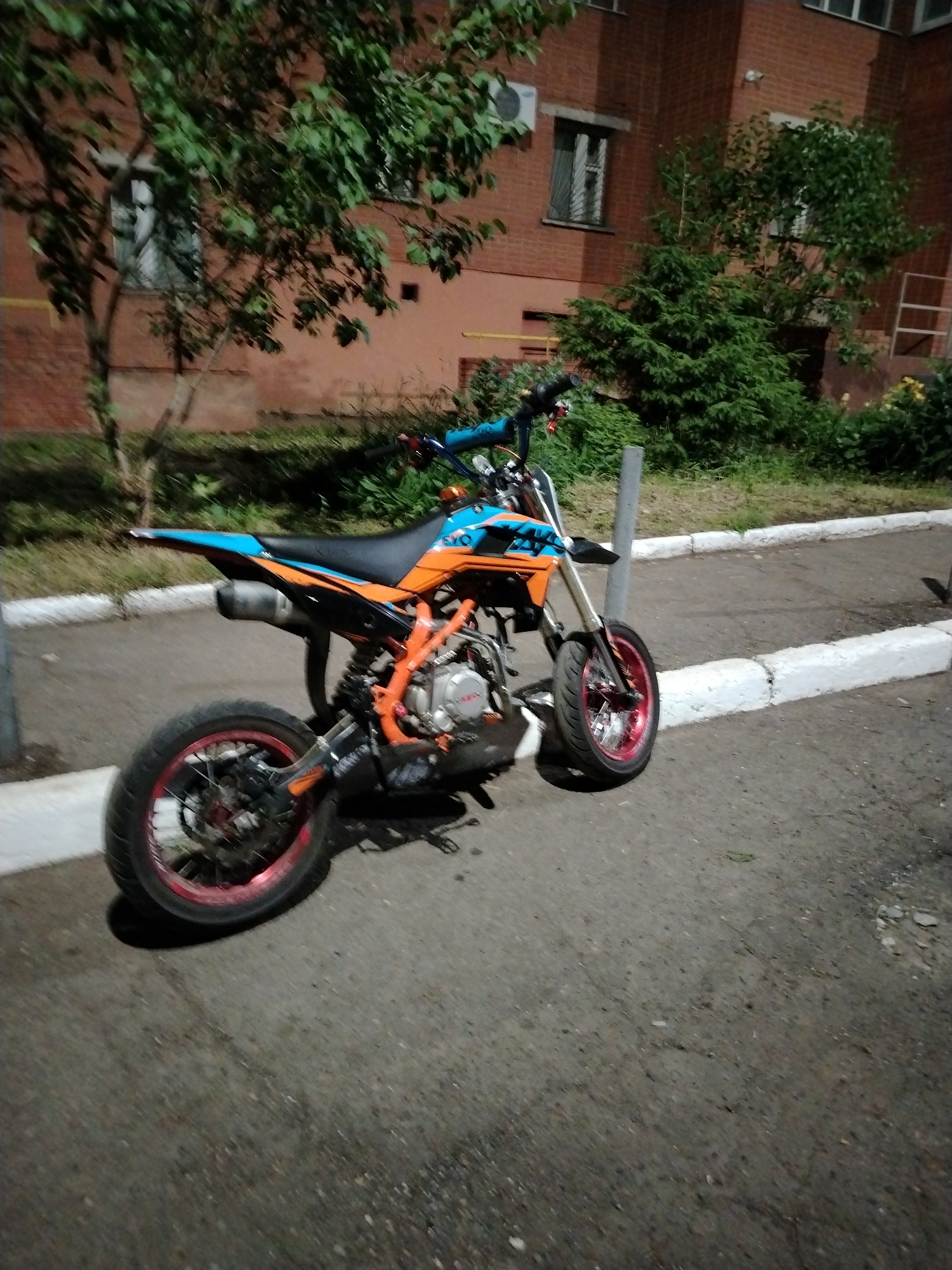 This image depicts a vibrant orange and blue pit bike, or small dirt bike, parked outdoors at dusk or night. The motorcycle features red wheel rims, black tires with disc brakes visible, and a black seat. The word "EVO" is clearly visible in blue letters on the side fairing, above the orange frame. The bike is resting on its kickstand on a paved asphalt surface.
In the background, a multi-story red brick apartment building is visible, consistent with residential architecture found in cities like Izhevsk, Russia. Several windows, some with white frames and one with an air conditioning unit, are set into the building's facade. Lush green trees and bushes, including a large deciduous tree and smaller conifers, occupy the area between the paved ground and the building. A white-painted concrete curb separates the asphalt from the vegetation. A grey cylindrical post stands near the motorcycle's front wheel.
The low light conditions suggest the photograph was taken during the evening or night. The scene is devoid of people, portraying a quiet, static moment in a residential or suburban setting. No specific event or activity is taking place beyond the motorcycle being parked. The weather appears clear.
