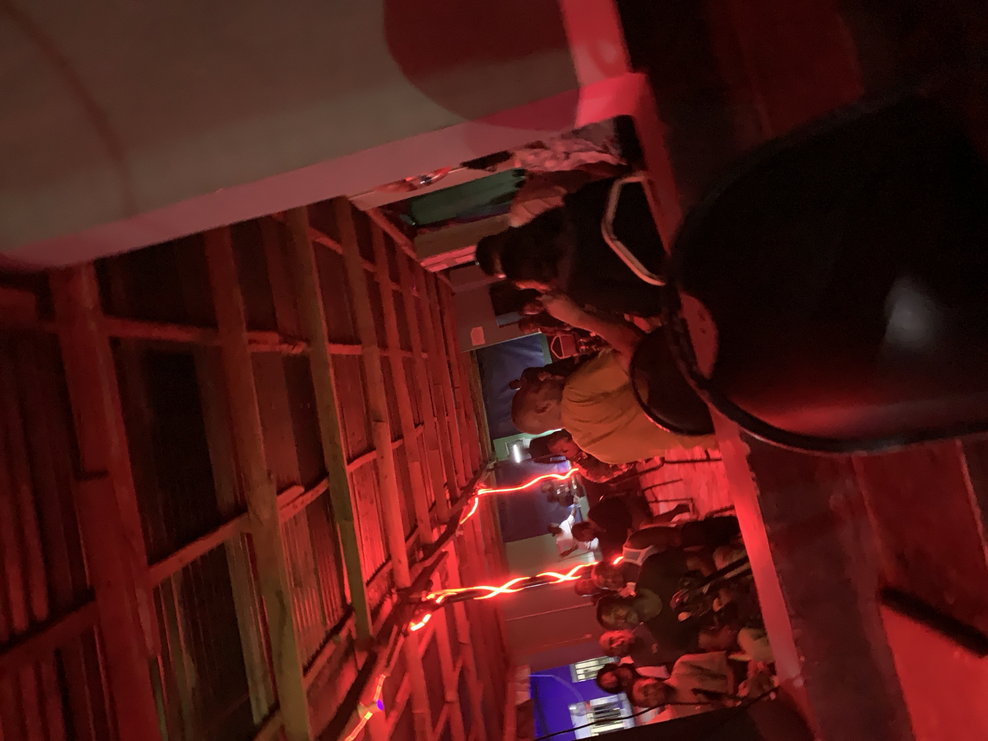 The image depicts a narrow alleyway in what appears to be a densely populated urban area, possibly in New Stadium, Nigeria, based on the query context. The alley is lit with red string lights, creating a dimly lit and vibrant atmosphere. The walls are constructed of corrugated metal and wooden beams, suggesting a makeshift or informal construction.
A crowd of people is gathered in the alley, seemingly for a social event or gathering. Individuals are seen wearing various clothing, including a yellow shirt and dark attire. There is a blurred object in the foreground that could be a speaker or part of a sound system.
The overall environment suggests a vibrant nightlife or community event taking place in a modest or possibly impoverished urban setting. The composition and lighting give the image a sense of depth and intimacy.
