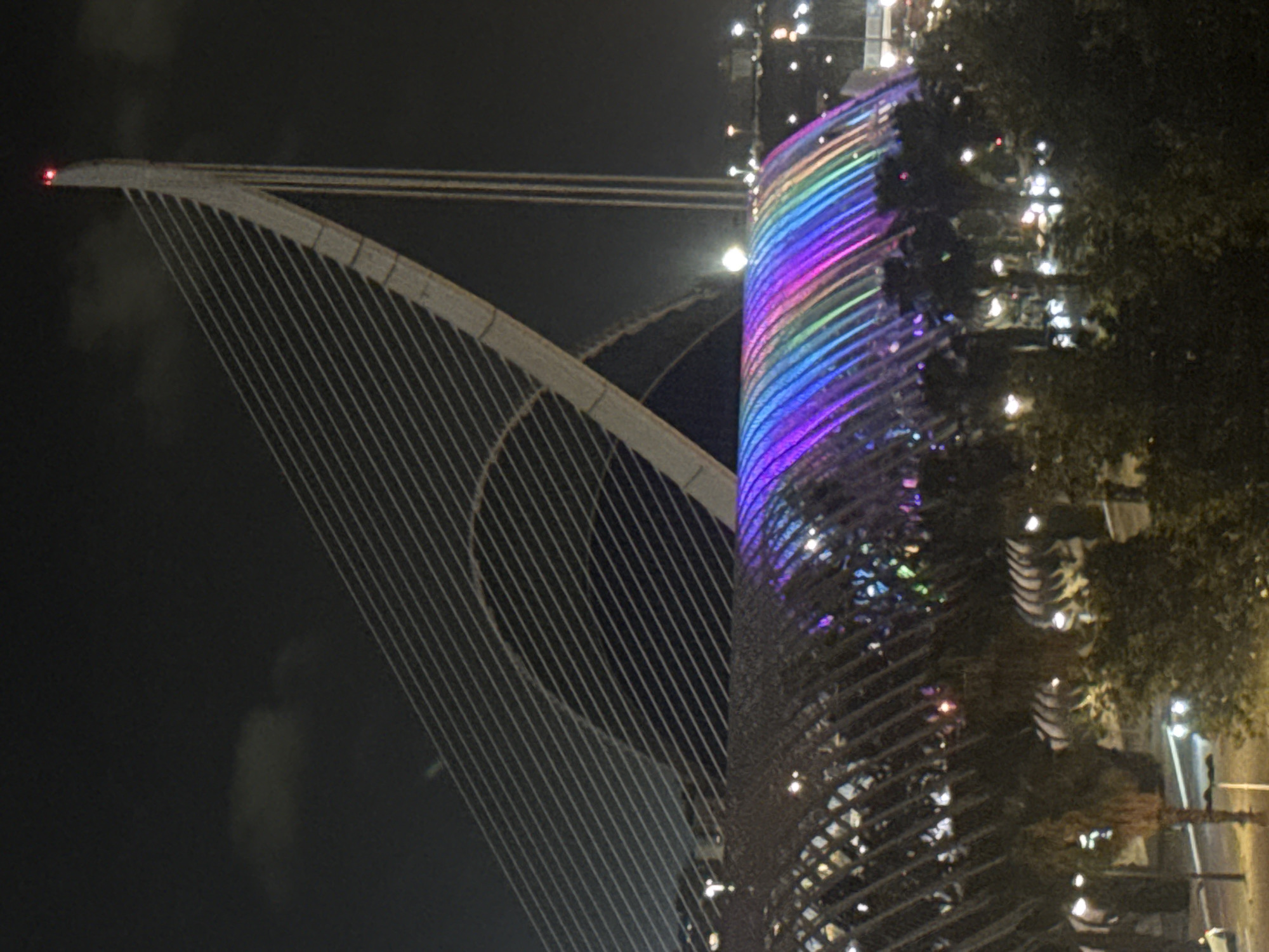 The image captures a nighttime scene within the City of Arts and Sciences complex in Valencia, Spain. In the upper right quadrant, the white, curvilinear pylon of the Assut de l'Or Bridge rises against a dark, overcast sky, with two small red lights visible at its apex. Multiple thin suspension cables extend diagonally downwards and leftward from the pylon. Below the bridge structure, the L'Hemisfèric building is centrally positioned, its large, arched roof structure glowing with a broad, horizontal spectrum of artificial light. The colors transition from vibrant blues and purples on the left to greens, yellows, and pinks on the right. Behind L'Hemisfèric, a larger, dark architectural arch is partially discernible. The lower half of the image is dominated by the dark forms of palm trees and other dense vegetation. Scattered bright white and yellow lights, consistent with streetlights and distant vehicle headlights/taillights, punctuate the dark foliage, indicating an active urban or park setting. No human figures are discernible within the frame.